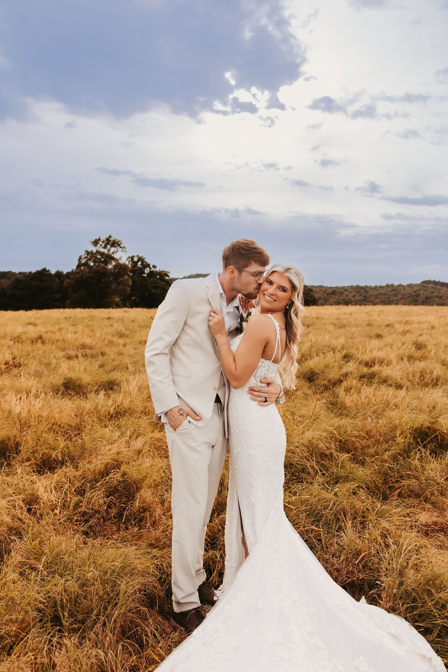 Bride and groom embrace in golden field, wearing white gown and beige suit, under a blue sky.