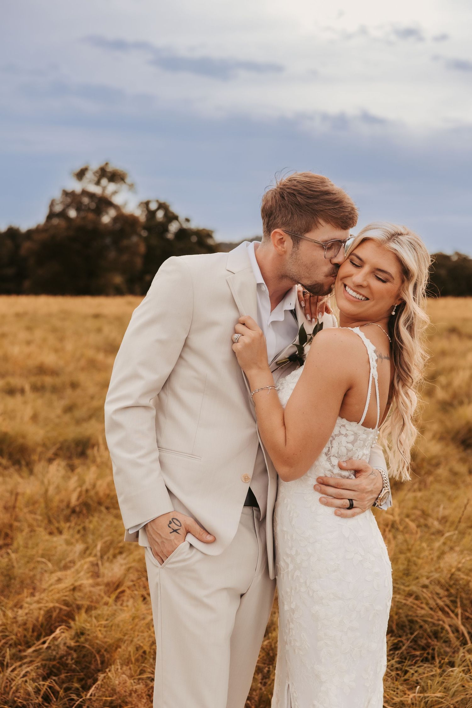 Groom in beige suit kissing bride on the cheek in a golden sunlit field.
