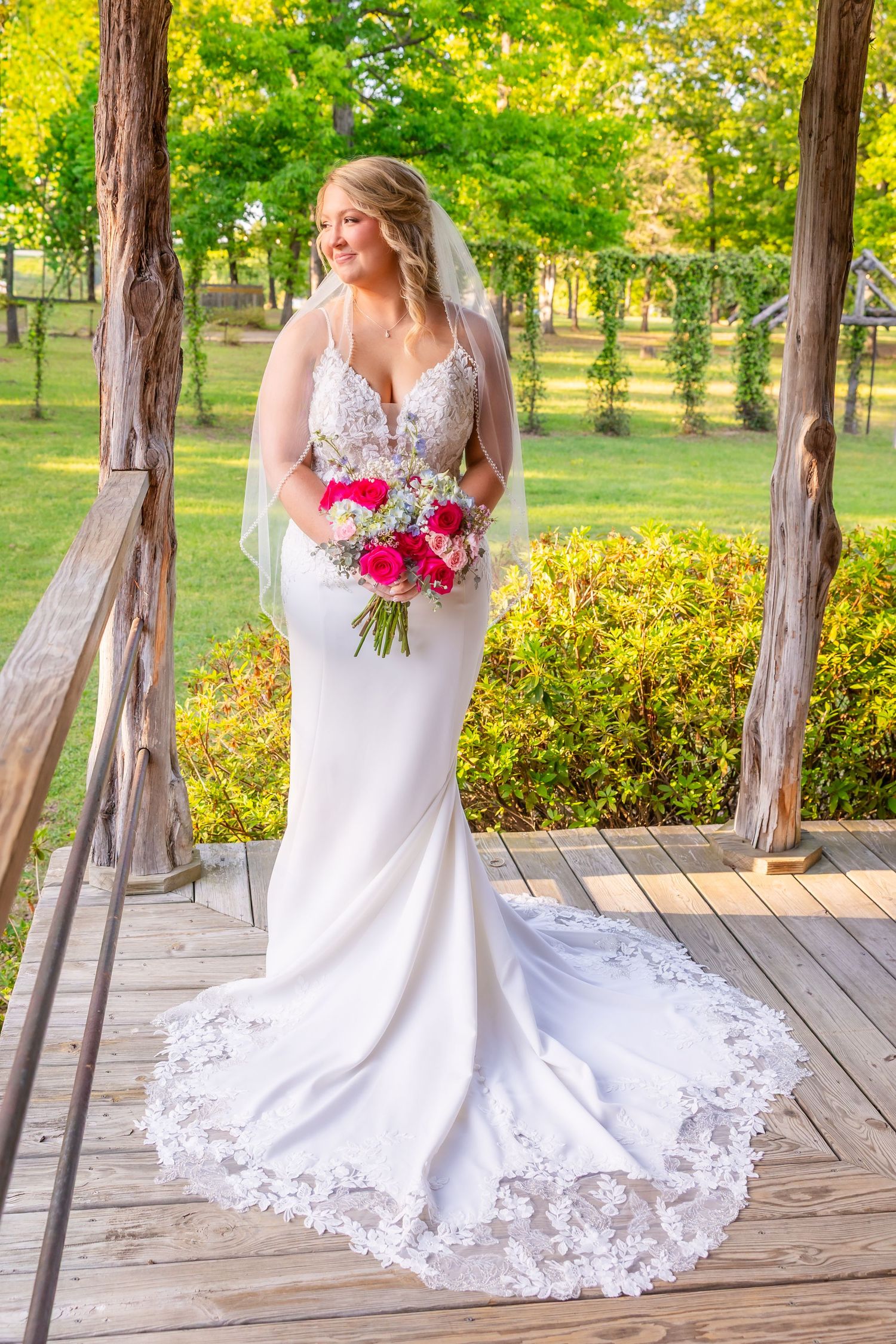 Bride in white lace mermaid gown with veil holds pink bouquet on wooden deck surrounded by lush greenery.