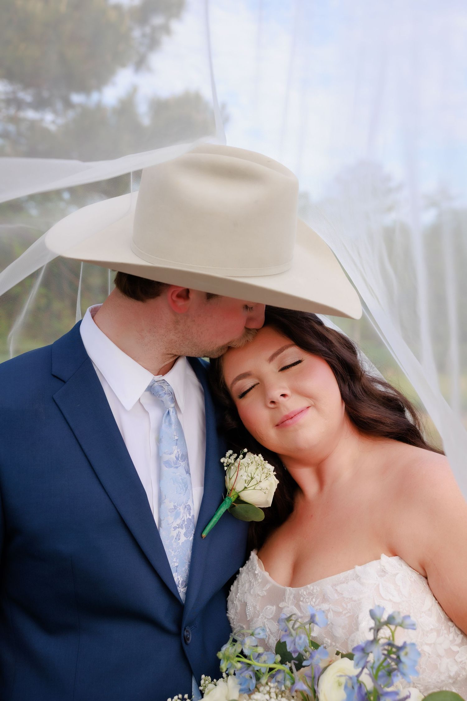 Groom in cowboy hat kisses bride on cheek as she smiles holding blue floral bouquet.