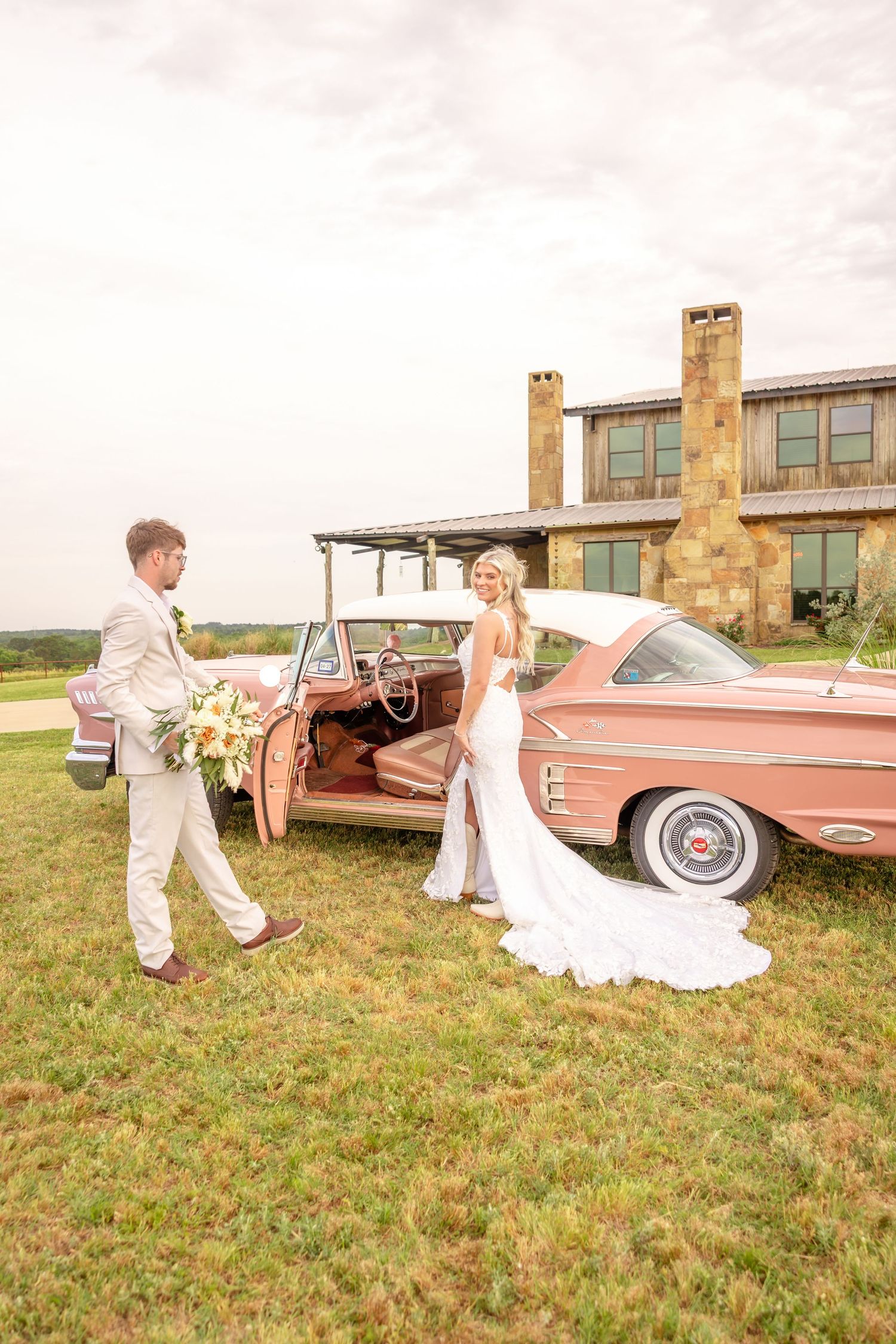 Groom opening door of a vintage pink car for his bride outside a rustic stone wedding venue.