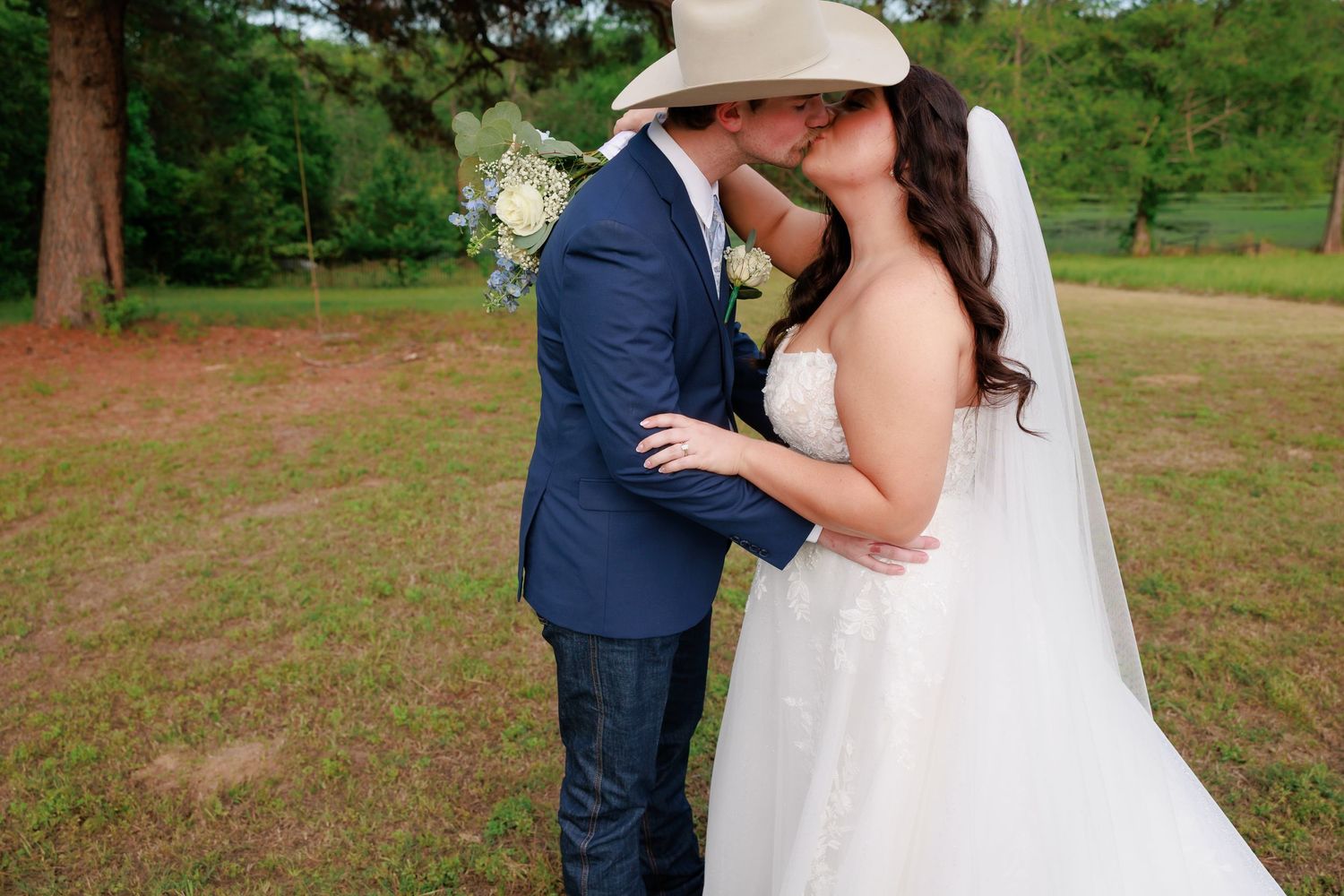 Bride and groom share a kiss outdoors with blue bouquet at rustic countryside wedding.