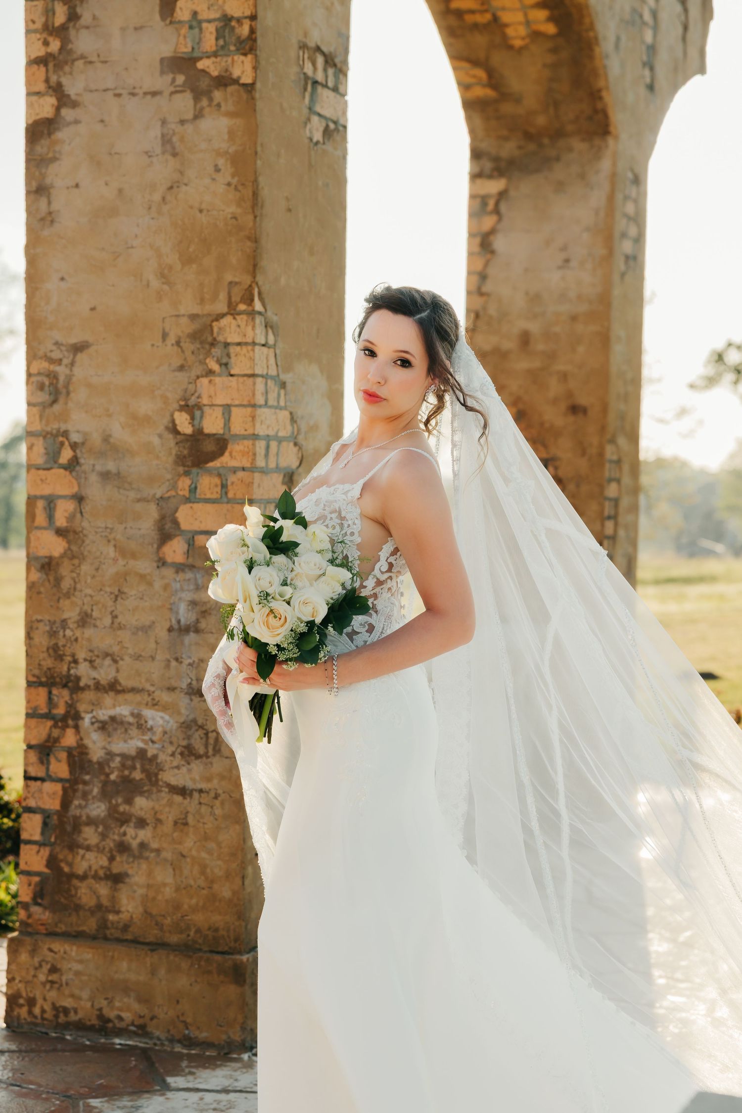 Bride in white lace gown and flowing veil holds white bouquet beside rustic stone archway columns.
