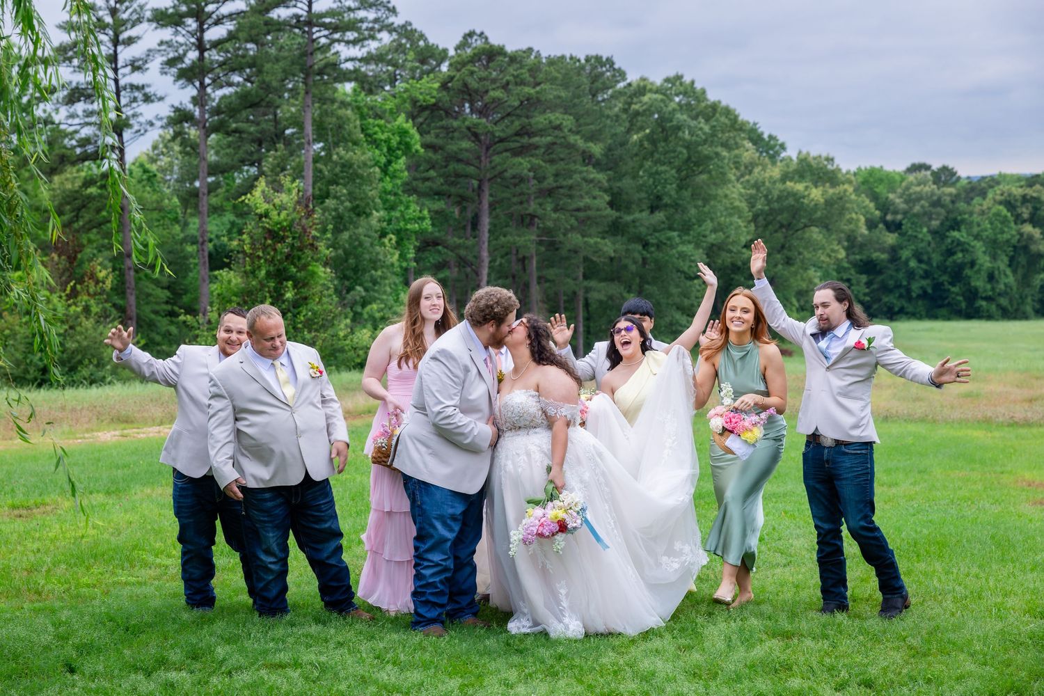 Wedding party celebrating outdoors on green grass with bride and groom kissing while bridal party cheers around them.
