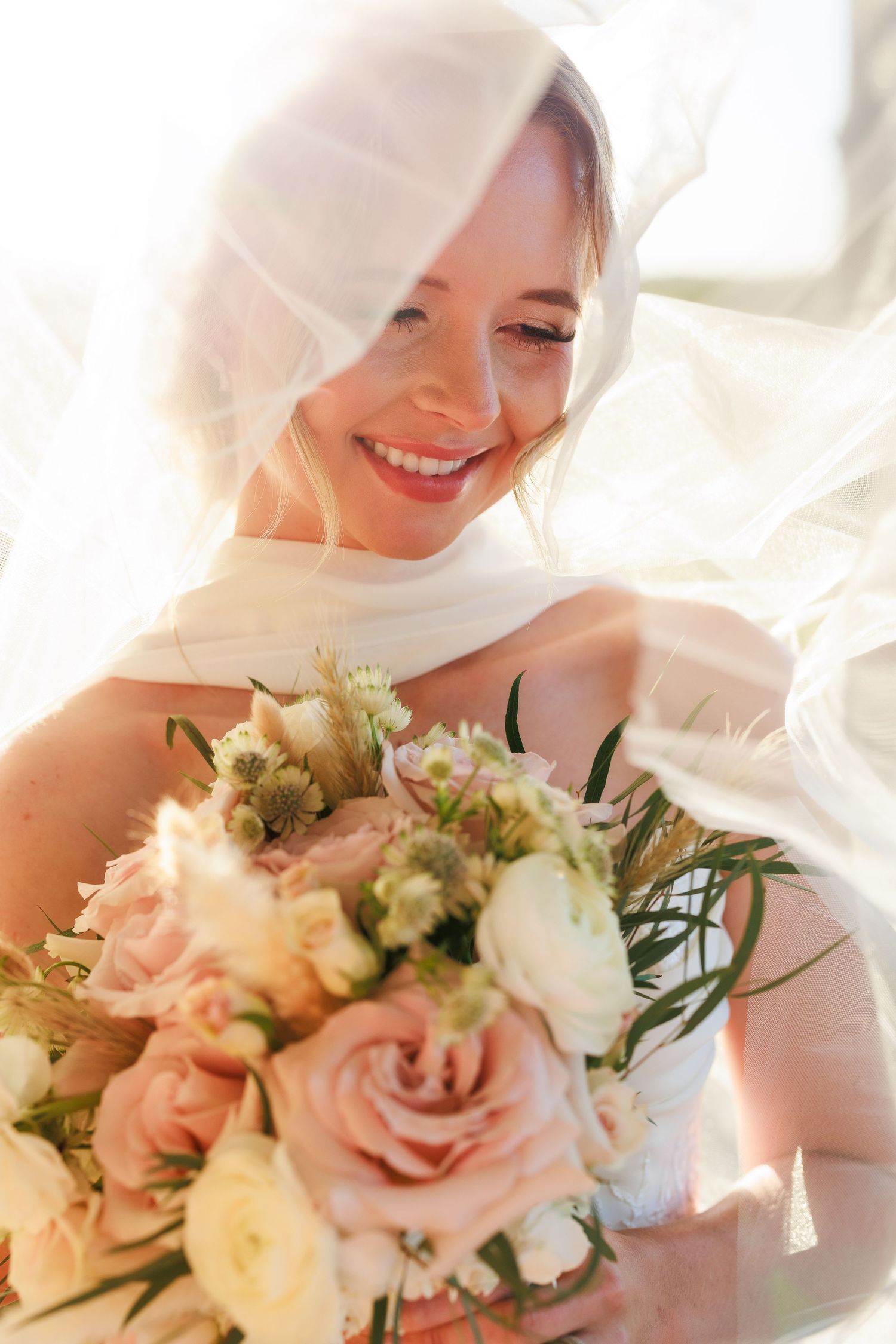 Radiant bride laughing under white veil while holding bouquet of blush roses and white flowers in sunlight.