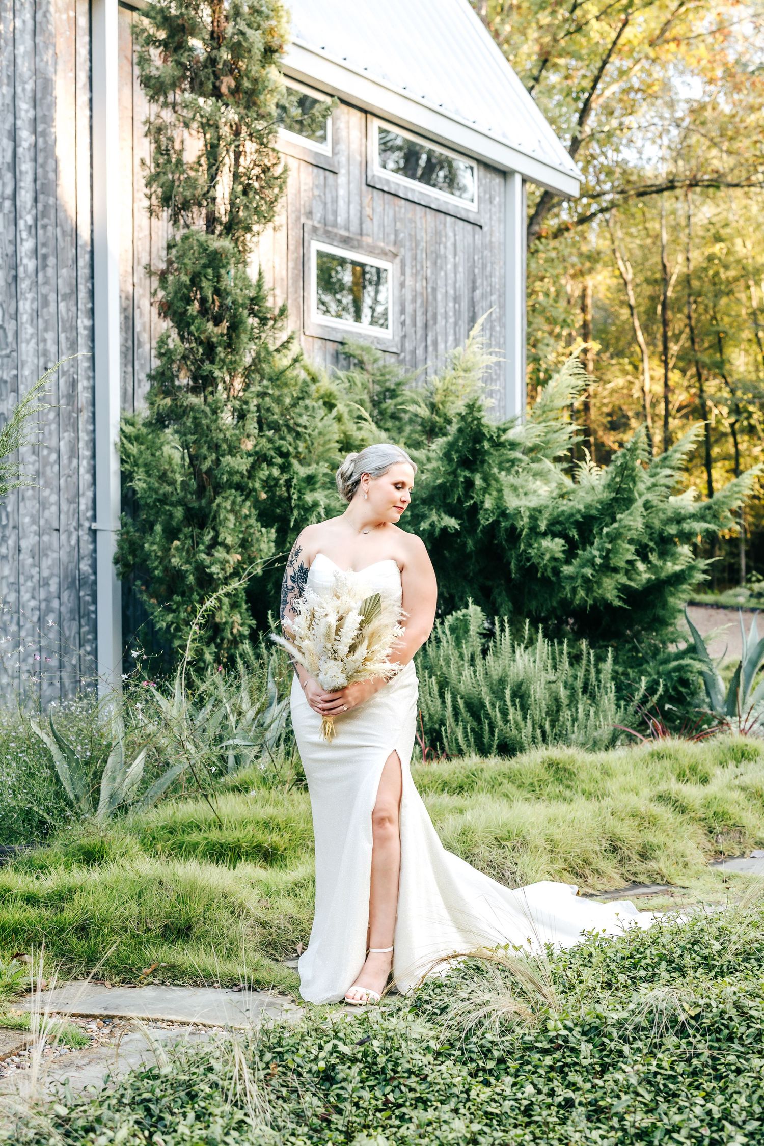 Bride in sleek white gown with slit holding bouquet beside rustic barn exterior.