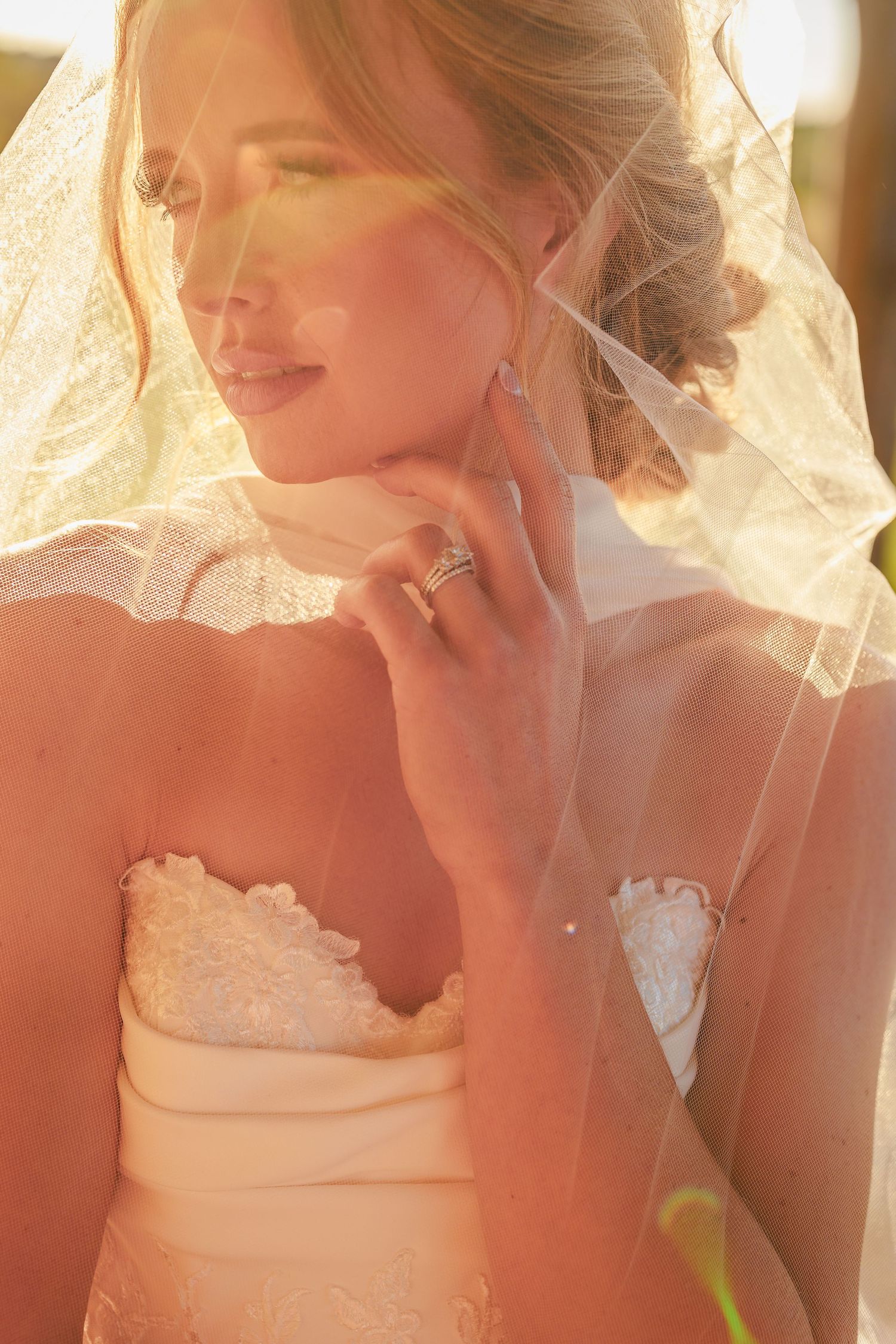 Bride in strapless lace gown with veil draped over her, touching her face gently in warm golden sunlight.