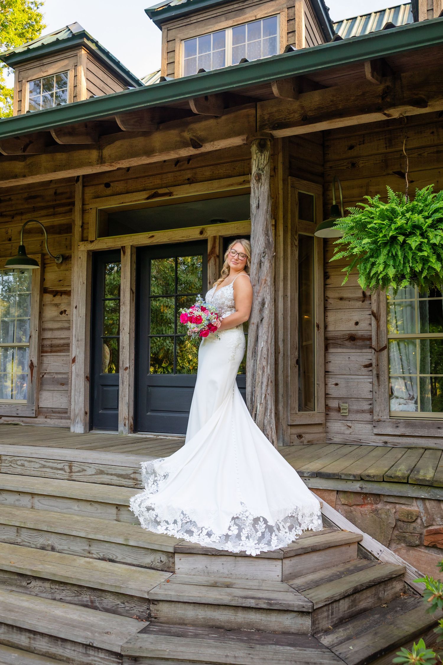 Bride in white mermaid gown holding pink bouquet poses on wooden porch steps of rustic log cabin venue.