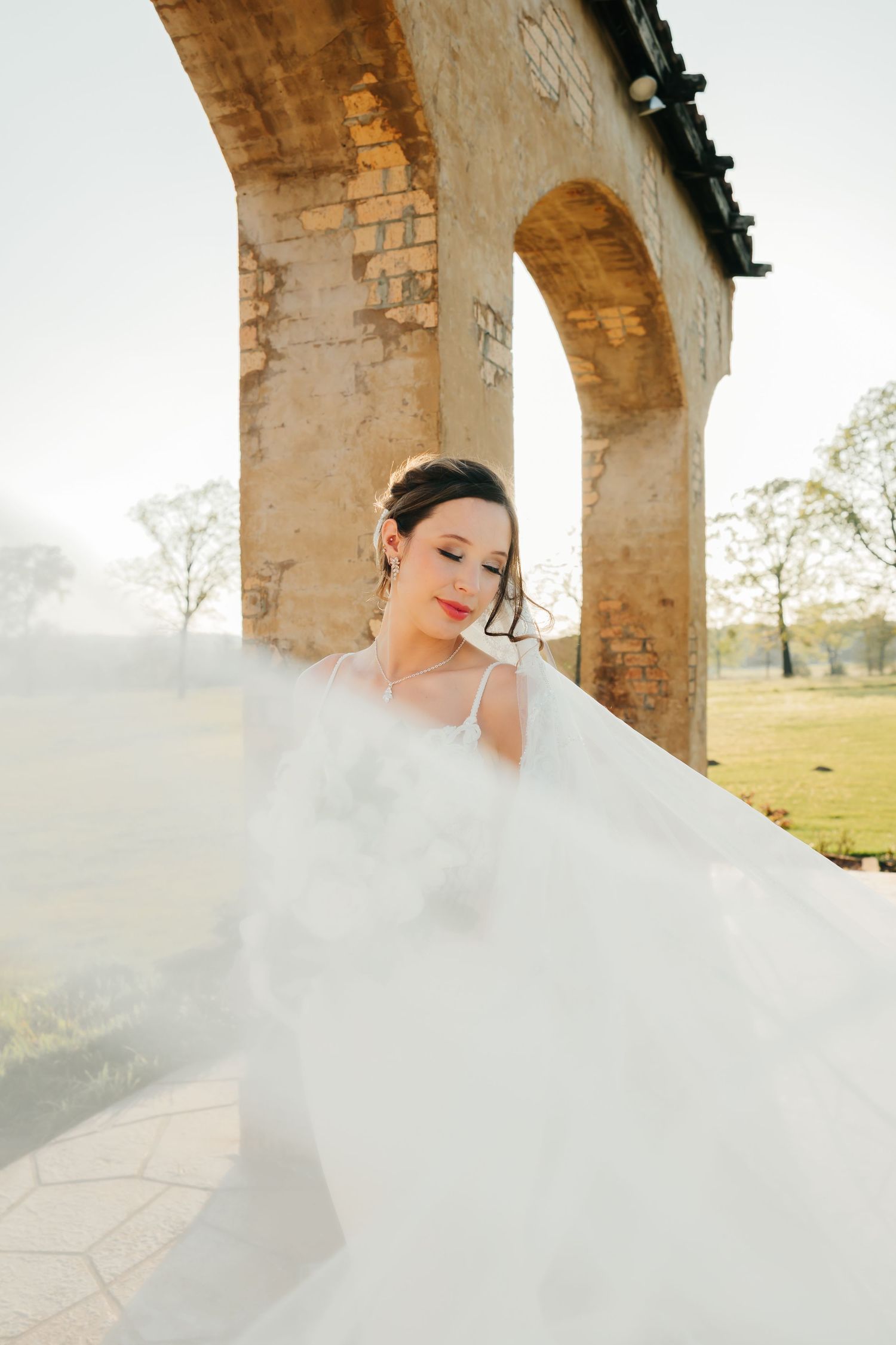 Bride in flowing white gown stands before rustic stone arches outdoors, veil billowing gracefully in soft natural light.