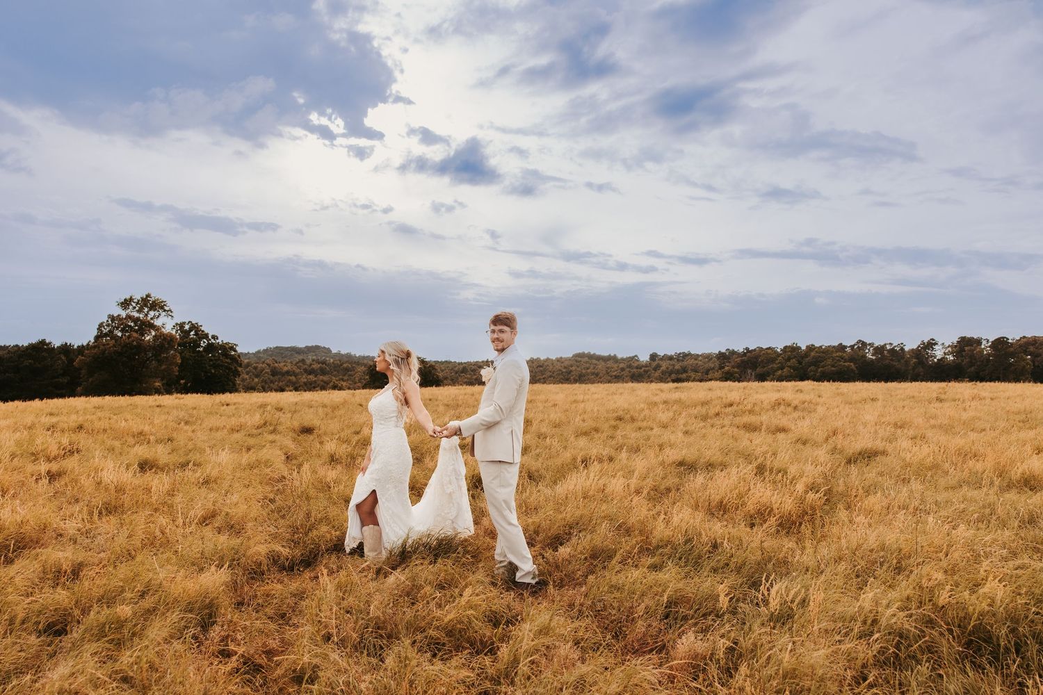 Bride and groom holding hands dancing in golden field under blue sky at outdoor country wedding.