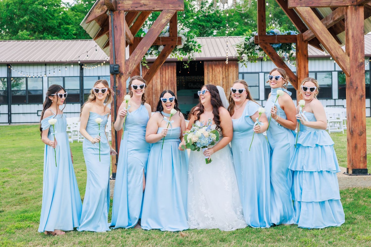 Bride poses with bridesmaids in light blue gowns and sunglasses under rustic wooden arch.