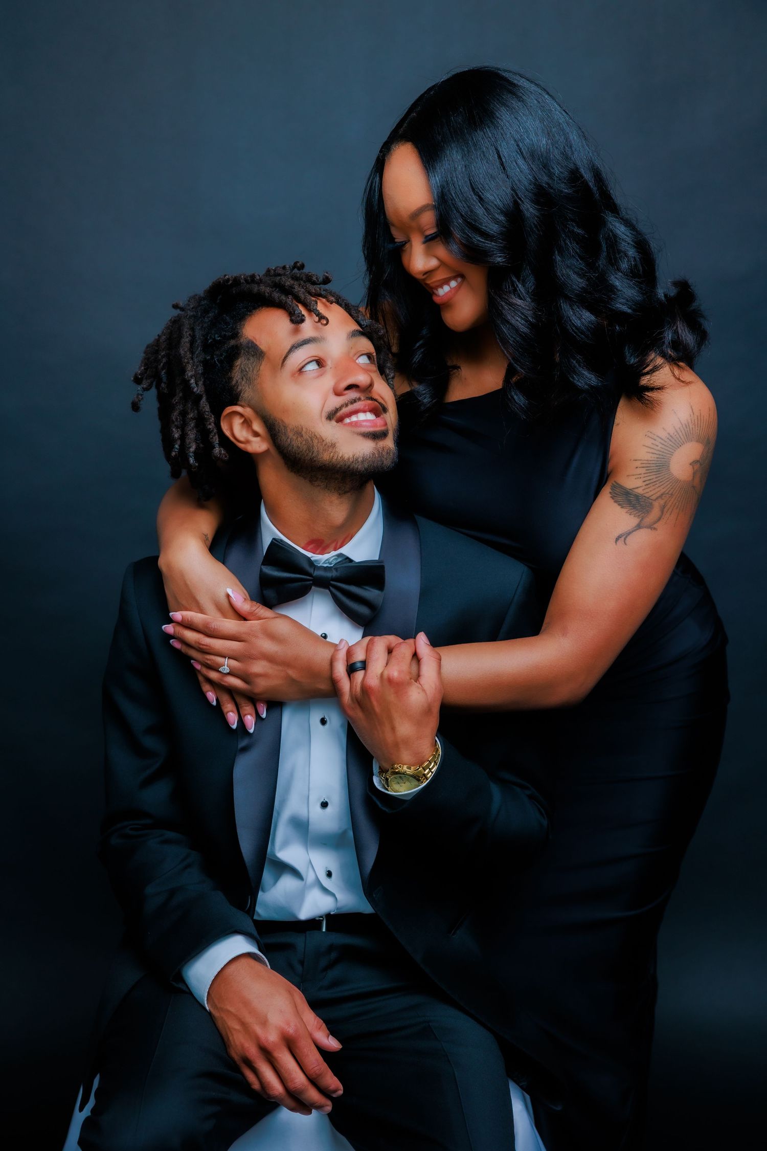 Couple in elegant black formal wear share a tender moment during studio engagement photo session.