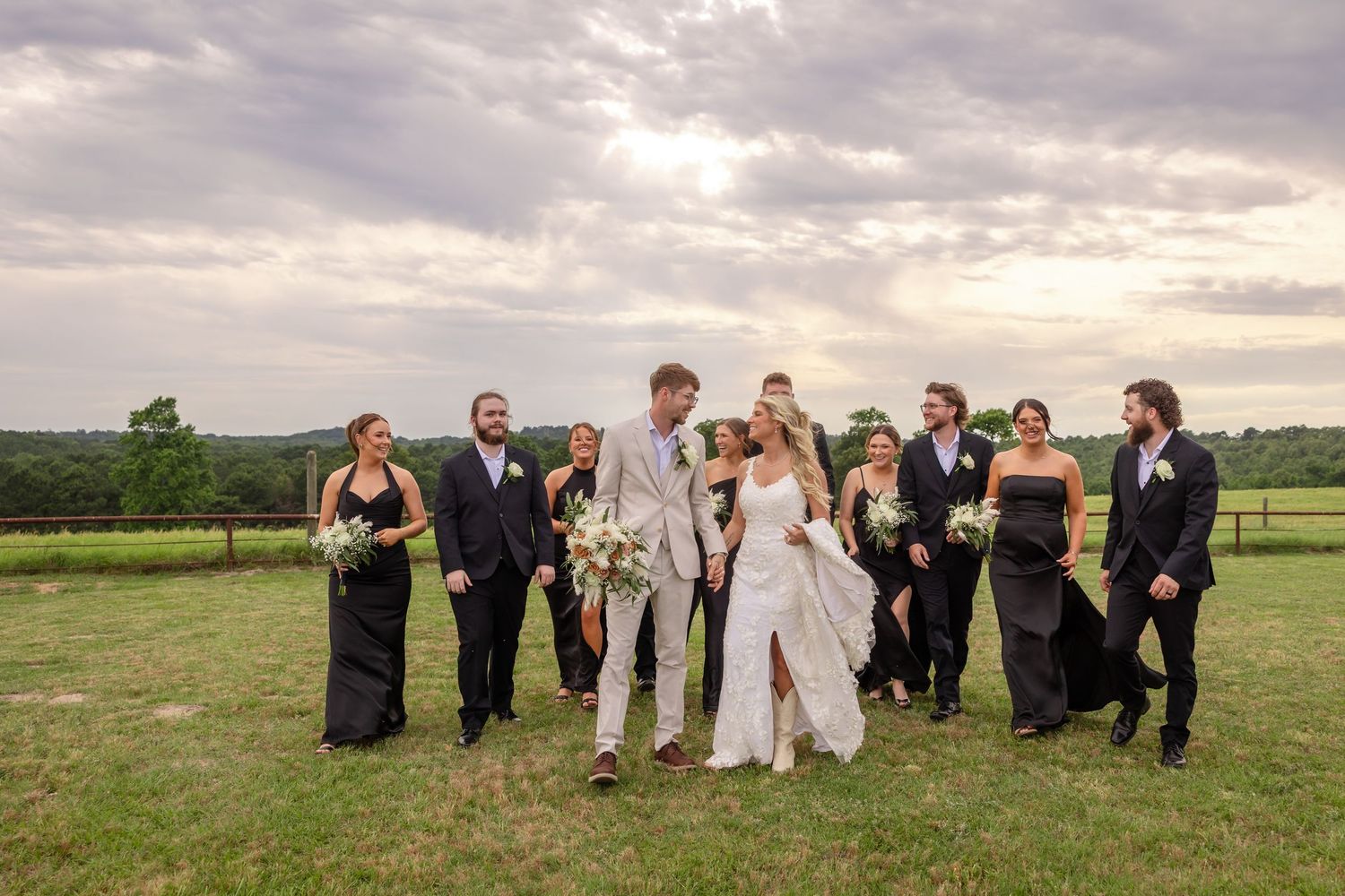 Wedding party in black and beige attire walking across a green field under a dramatic cloudy sky.