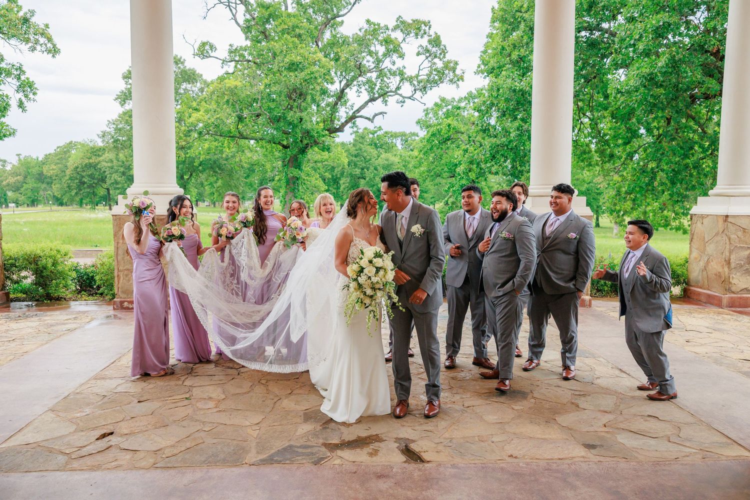 Wedding party poses under stone columns, bridesmaids in lavender gowns and groomsmen in gray suits surround the bride and groom outdoors.