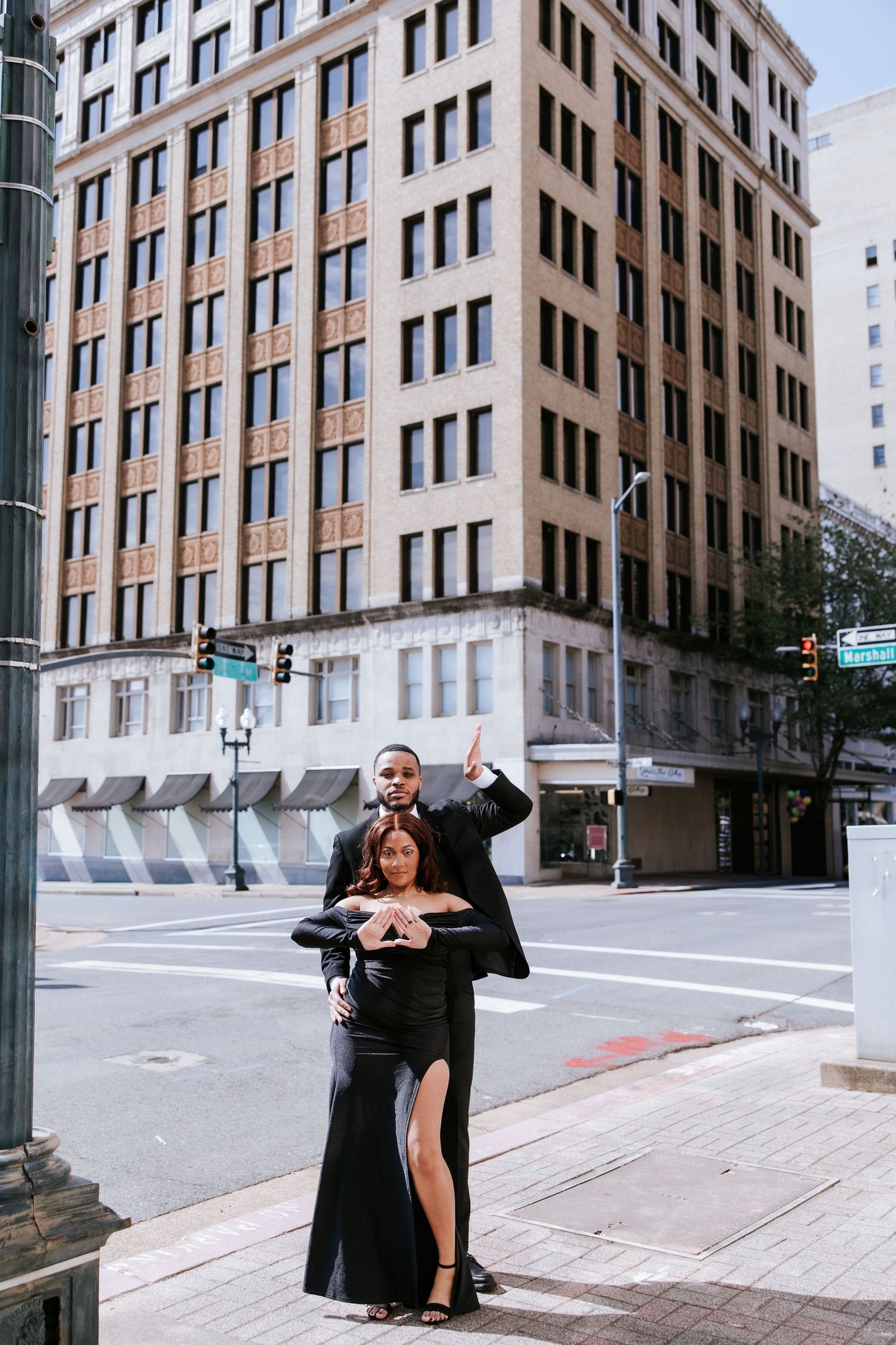 Couple in black formal wear posing on urban street corner with multi-story building in the background.