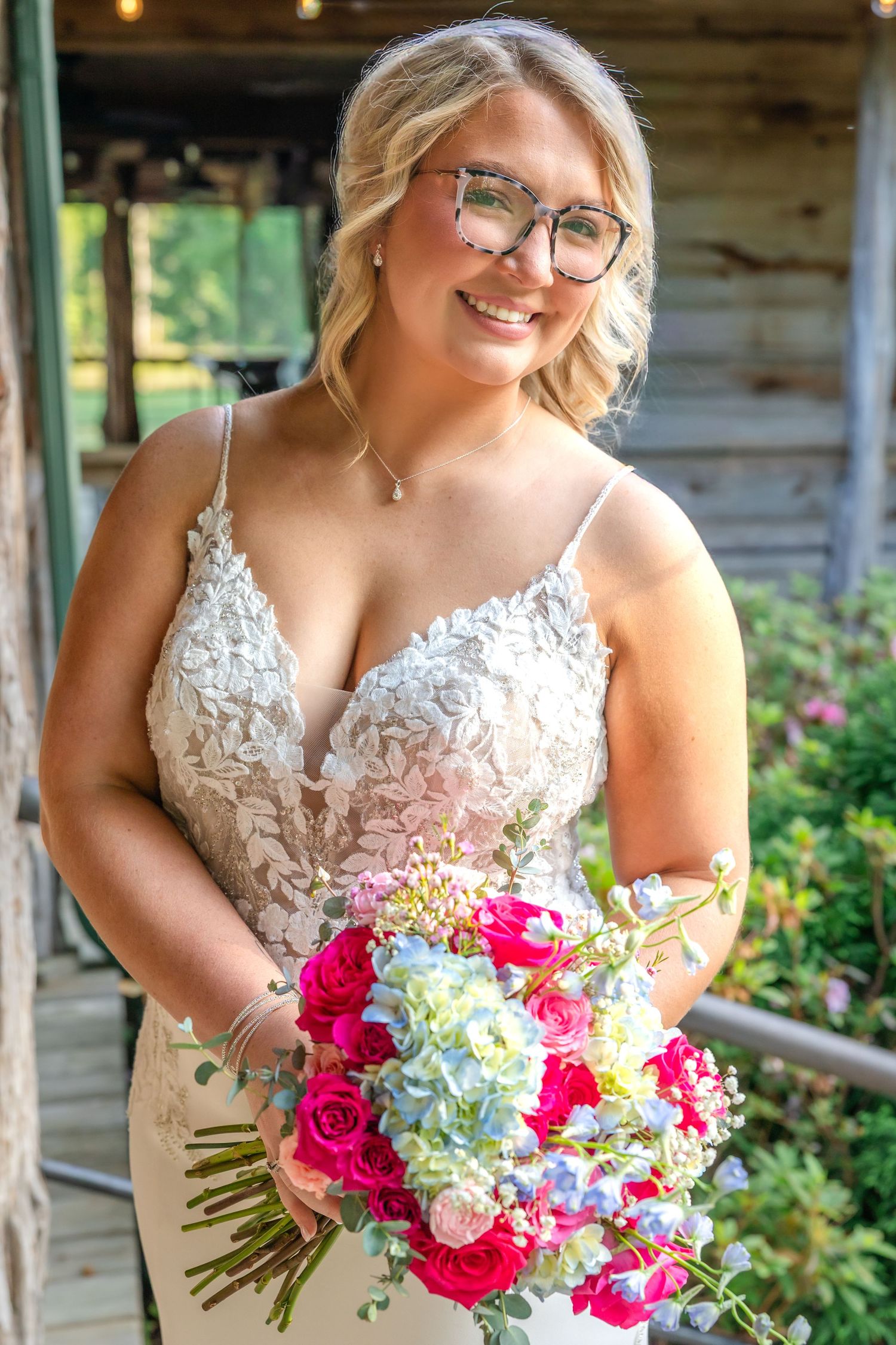 Smiling bride in lace wedding gown holding colorful bouquet of pink roses and hydrangeas, wearing glasses outdoors.
