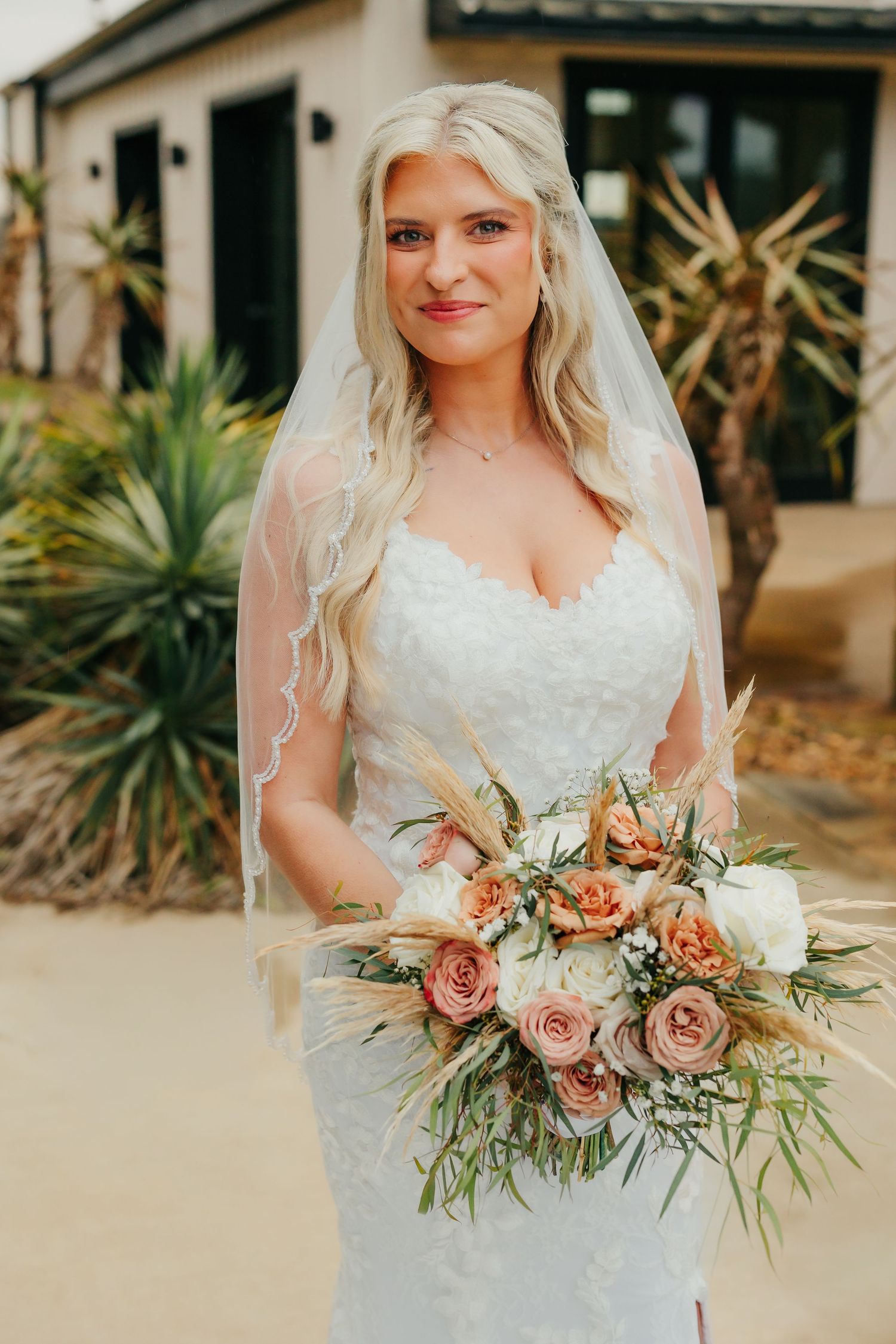 Bride in lace wedding dress and veil holds boho bouquet of peach roses and pampas grass outdoors.