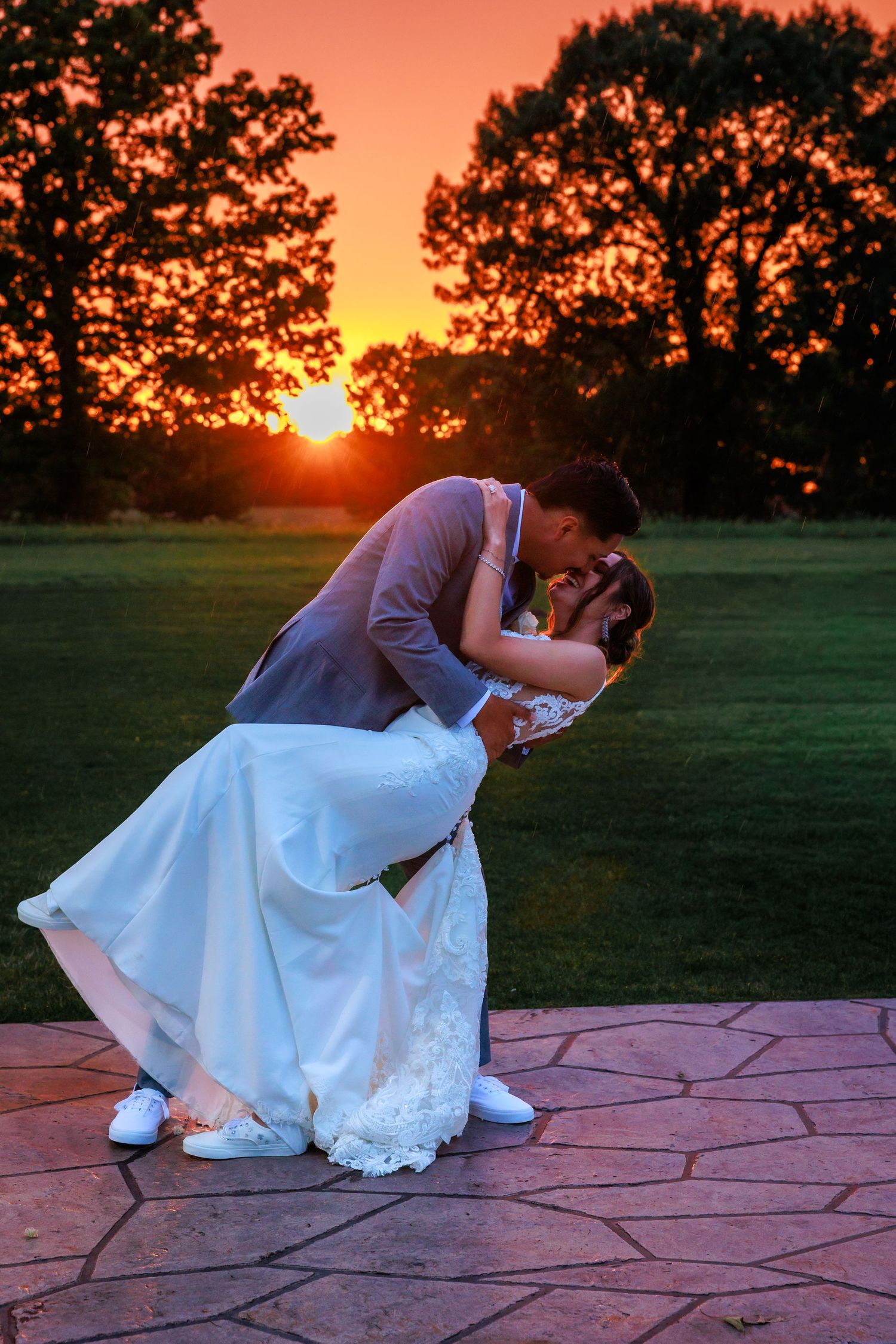Couple sharing a romantic dip kiss at sunset on a stone patio surrounded by trees.