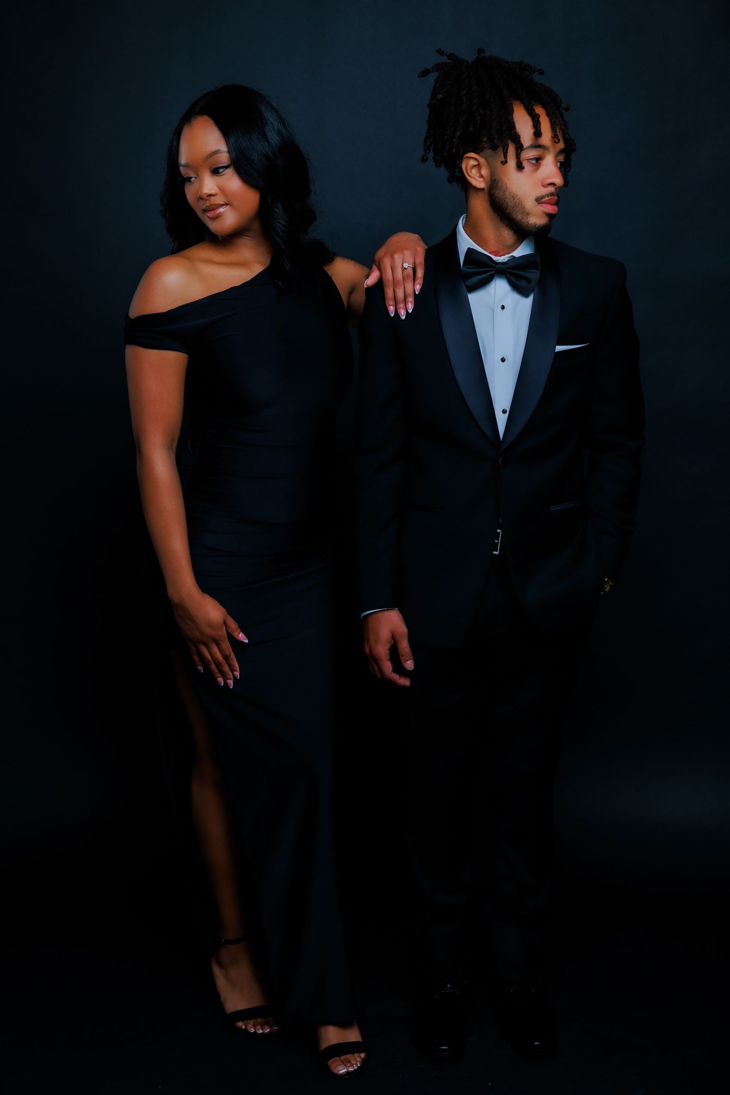 Stylish couple in coordinating black formal outfits pose together during dark studio photo session.