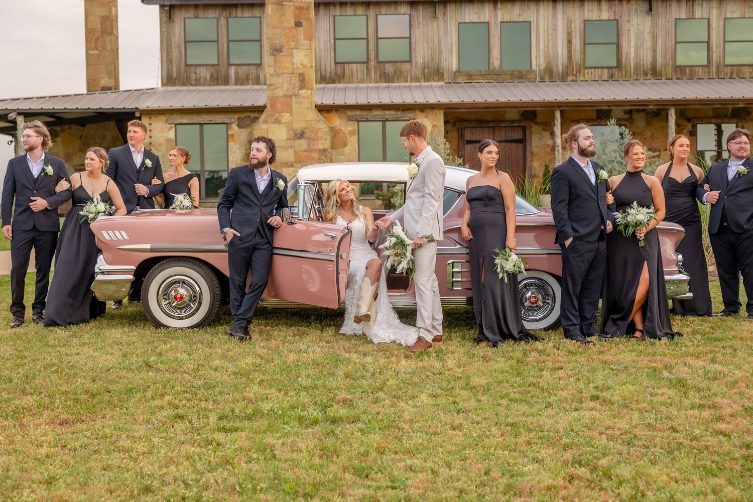 Bridal party posing with a vintage pink car in front of a rustic stone wedding venue.