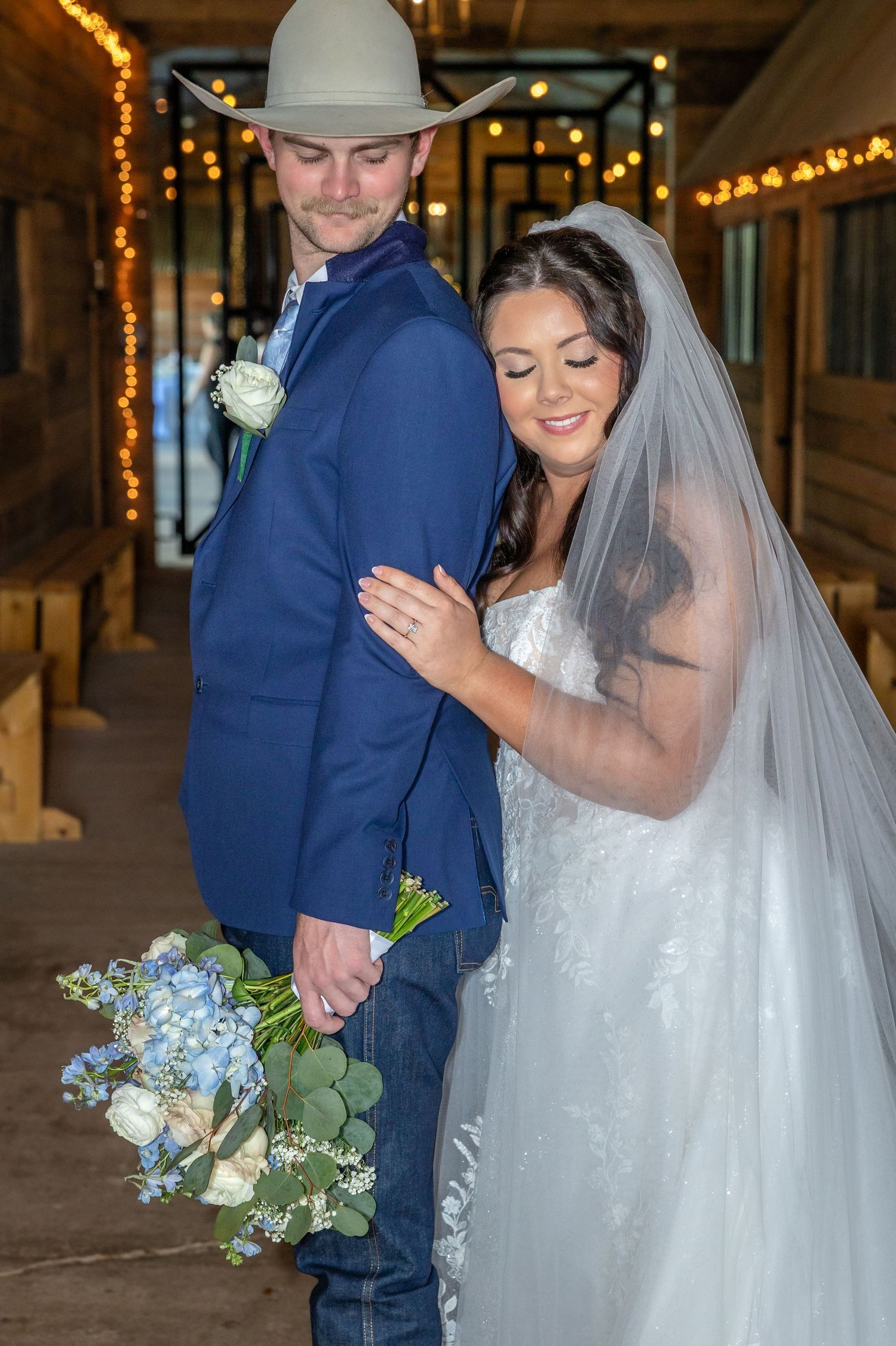 Bride hugs groom in blue suit and cowboy hat inside warmly lit rustic barn venue.