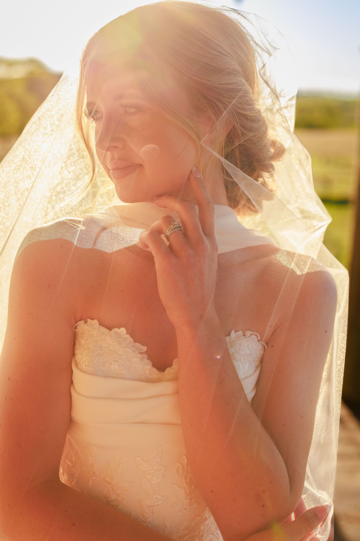 Bride in lace wedding dress and veil glowing in golden sunlight, smiling softly outdoors.