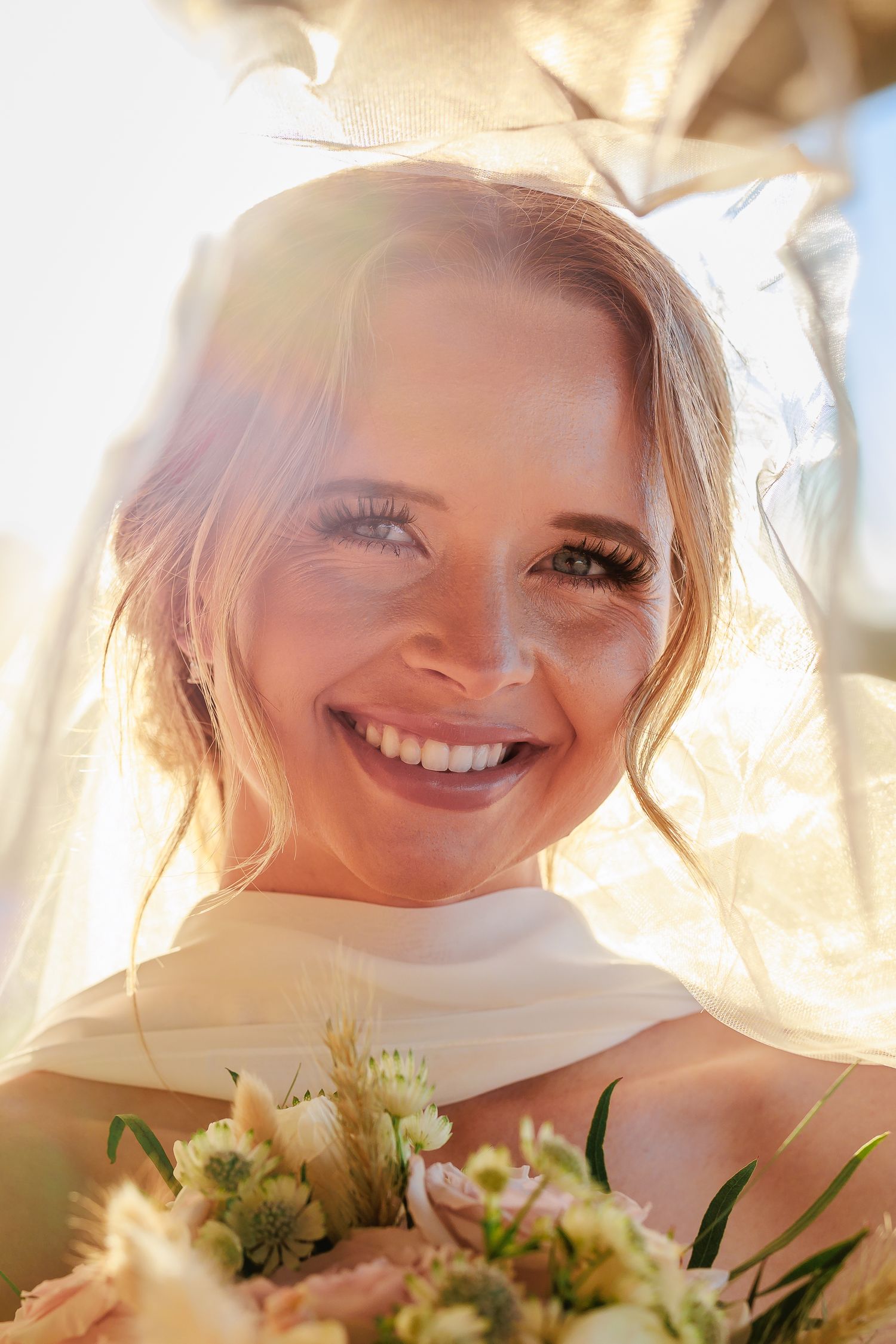 Smiling bride under flowing veil holding bouquet of pastel flowers, bathed in bright golden sunlight.