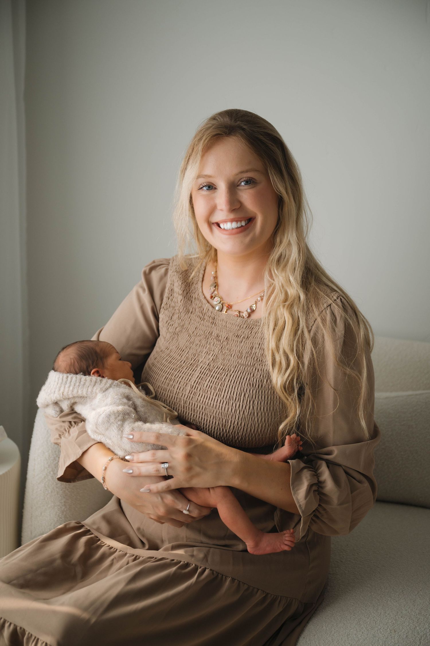Smiling blonde mother in beige dress holding sleeping newborn baby while sitting on a couch.