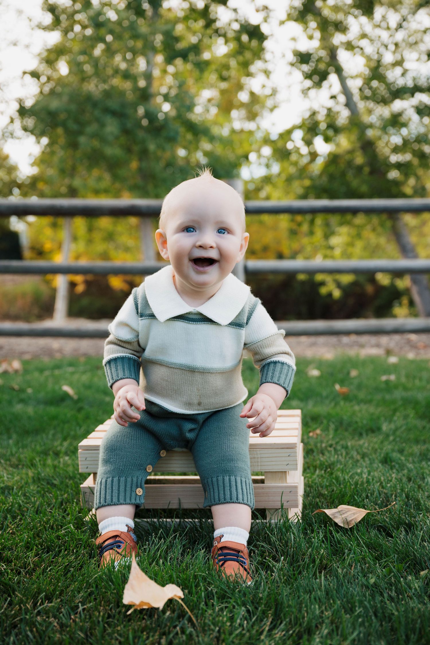 Baby in blue and white outfit sits smiling on wooden bench outdoors.