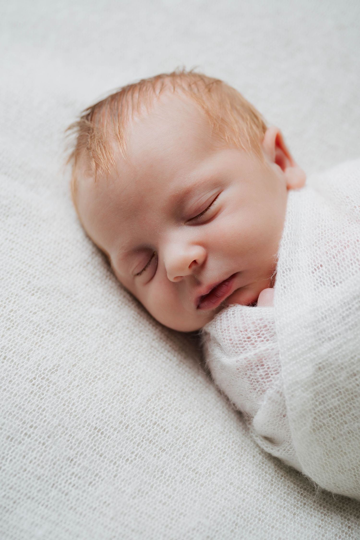 Close-up profile view of sleeping newborn wrapped in white knit blanket against light background.
