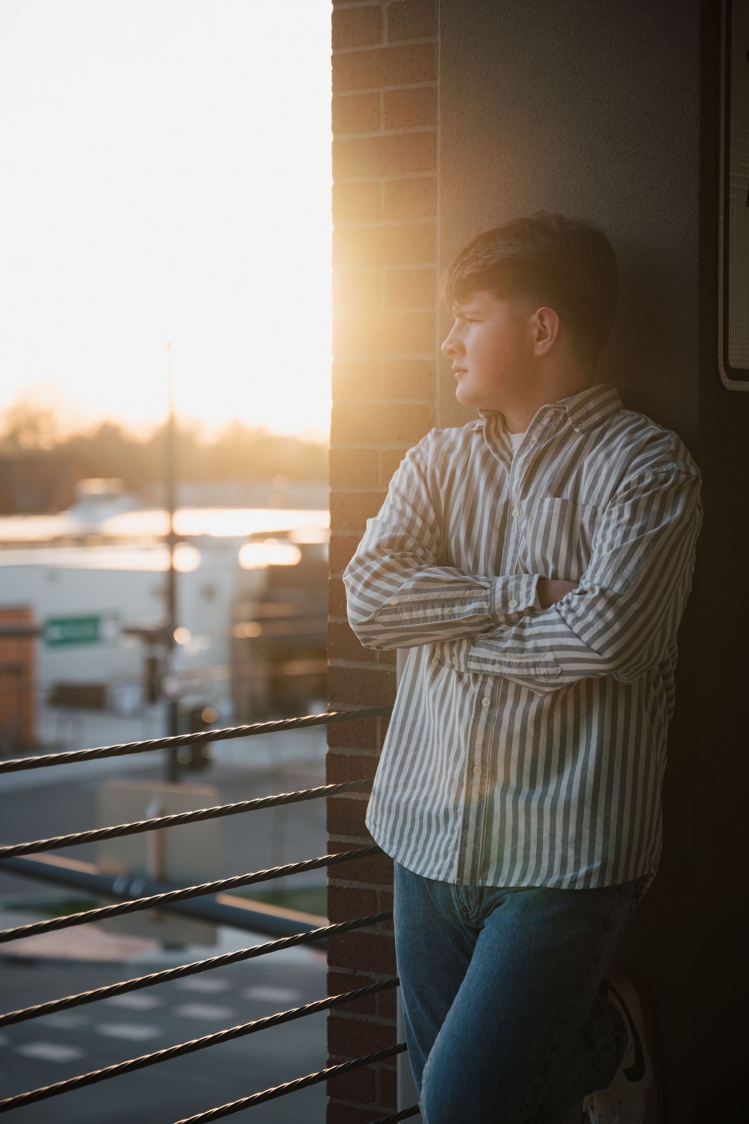 Sunset portrait by railing showing contemplative pose in striped shirt and jeans.