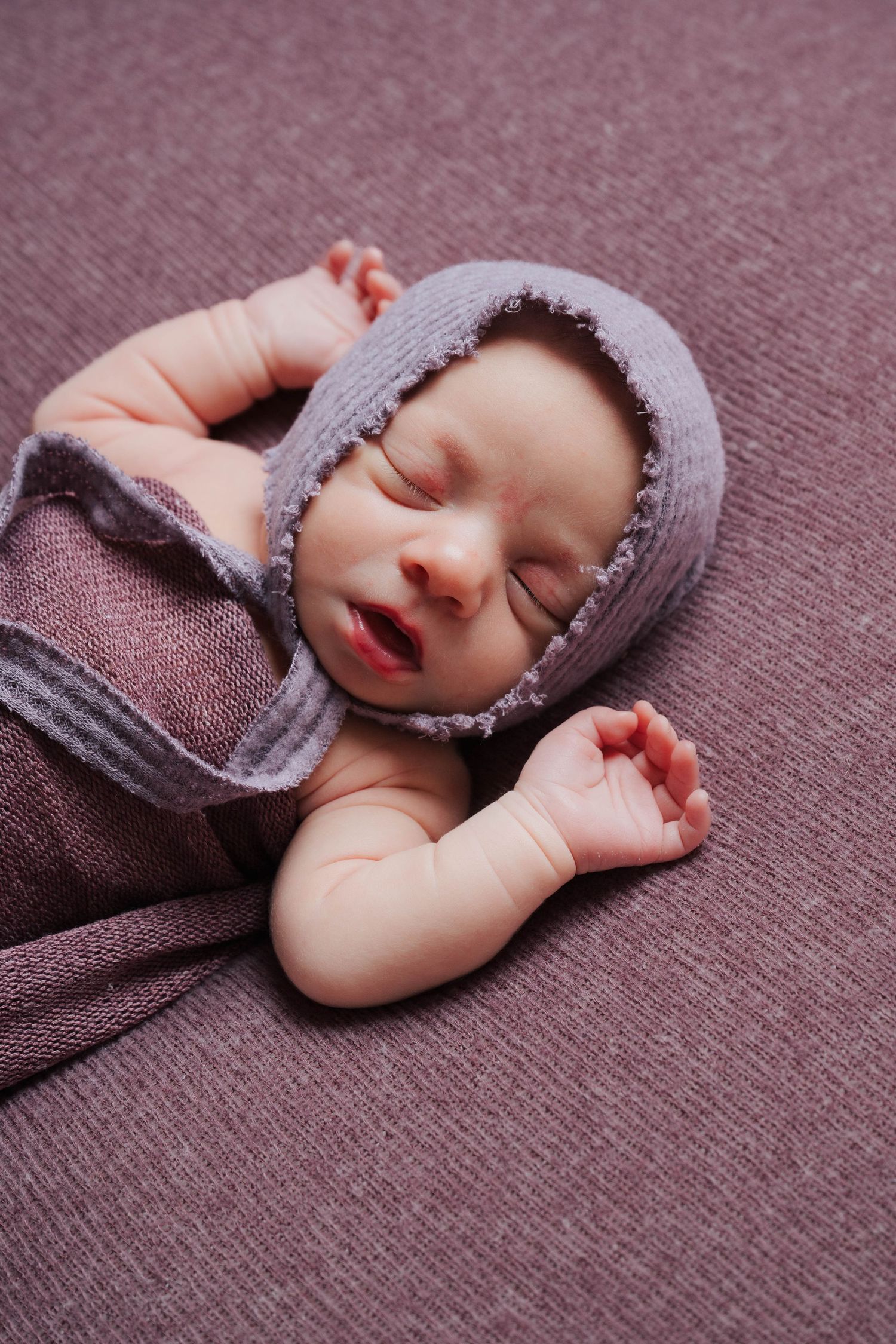 Peaceful newborn baby sleeping in purple knit wrap on textured purple surface.