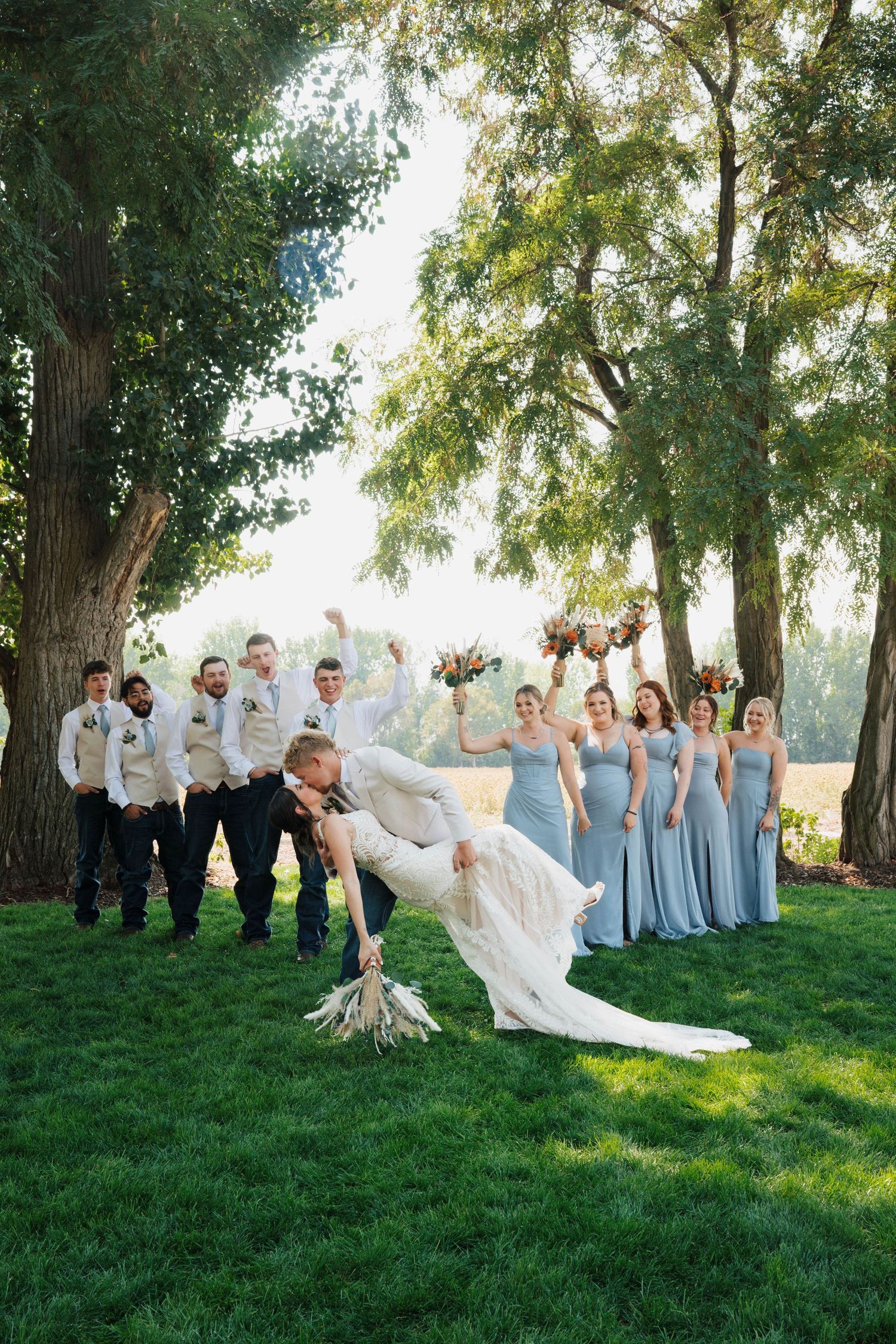 Bride and groom share a romantic dip kiss while wedding party cheers behind them on lush green lawn under tall trees at Deer Flat Ranch