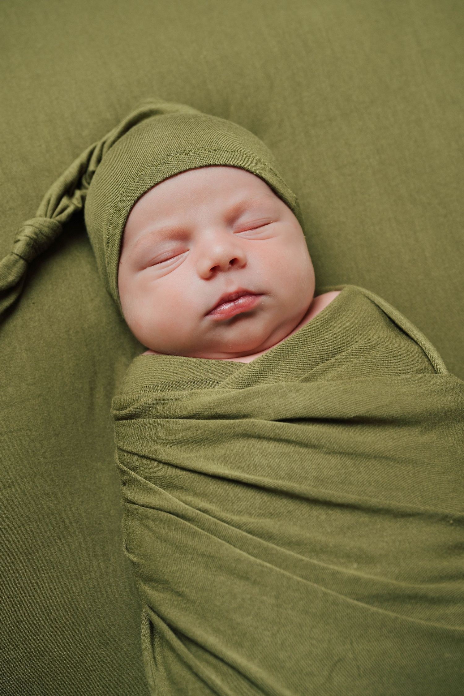 Sleeping newborn wrapped in olive green swaddle against matching background.