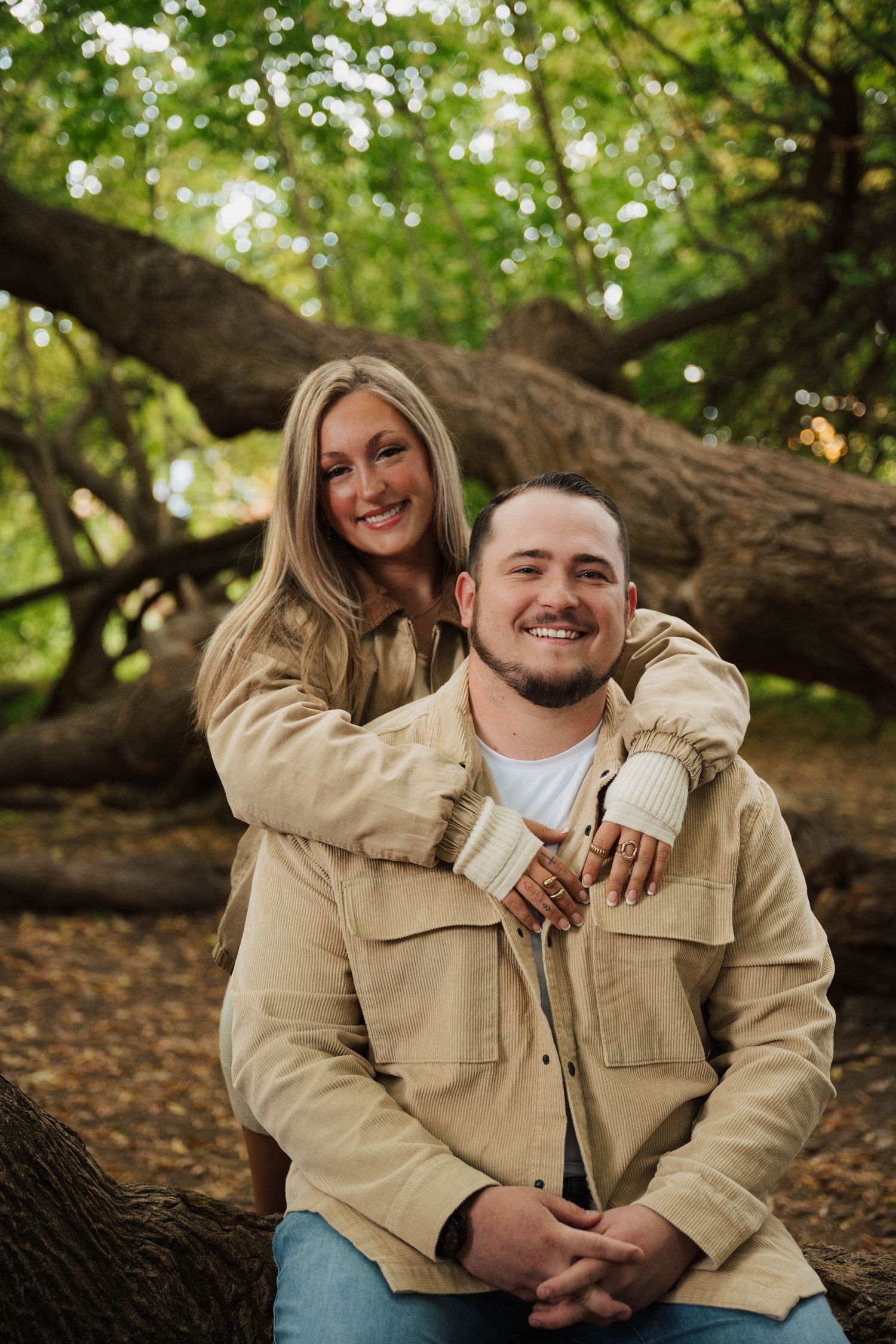 A couple sits together on a fallen tree branch in a lush green forest setting.