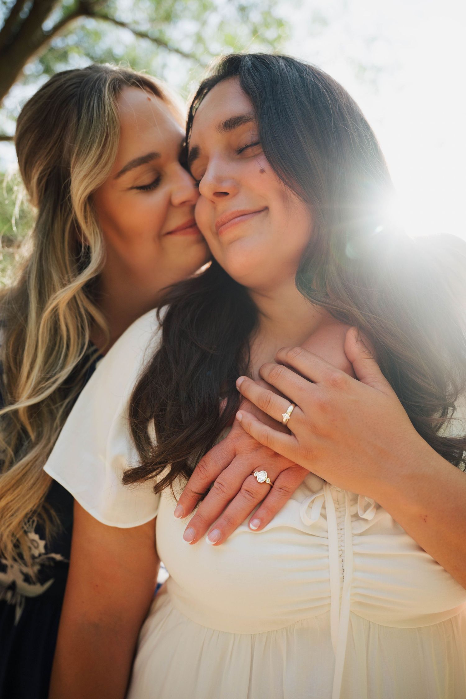 Two women sharing a tender moment outdoors, one kissing the other's cheek, displaying an engagement ring in sunlight.