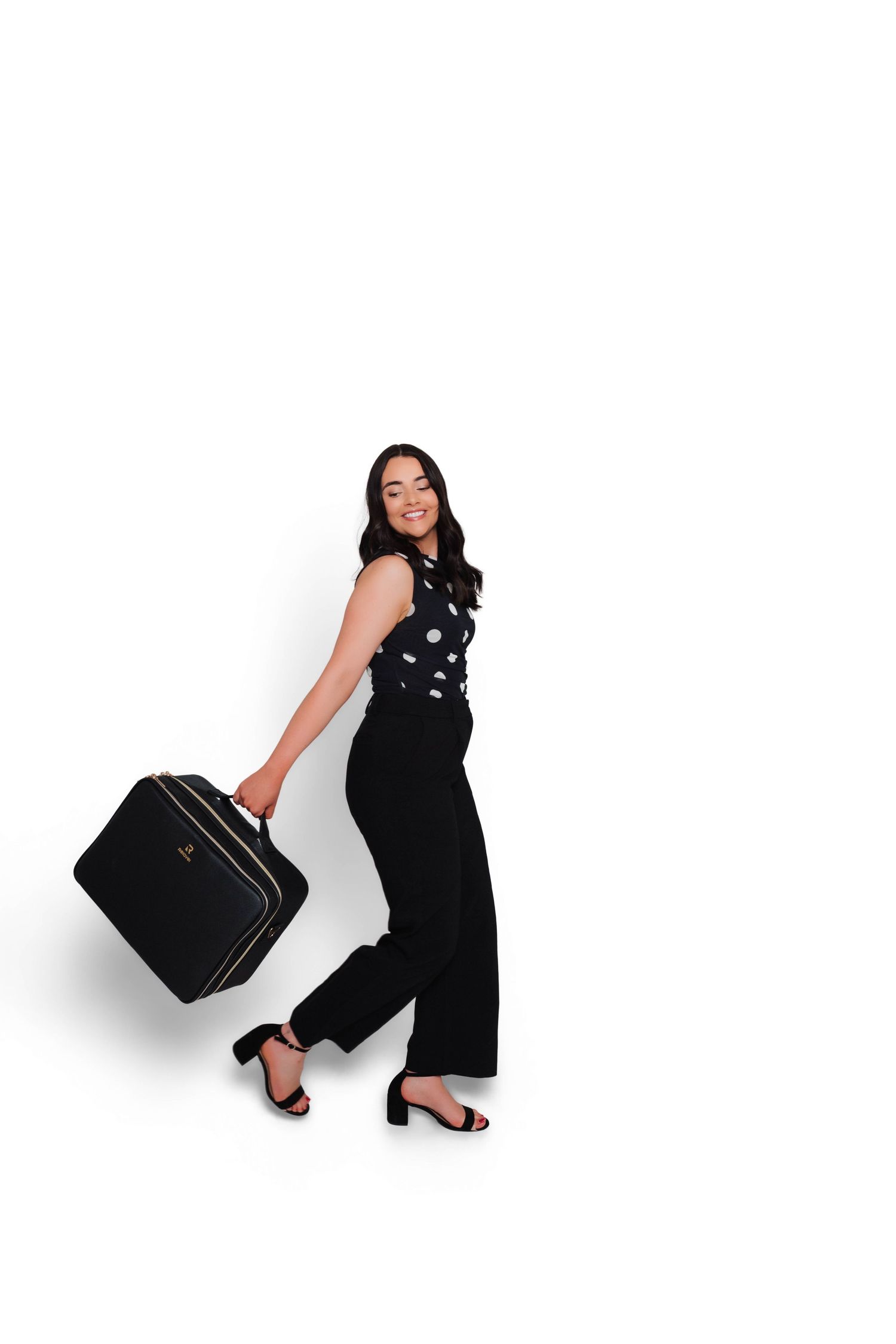Woman in black polka dot top and wide-leg pants carrying a black suitcase, walking confidently on white background.