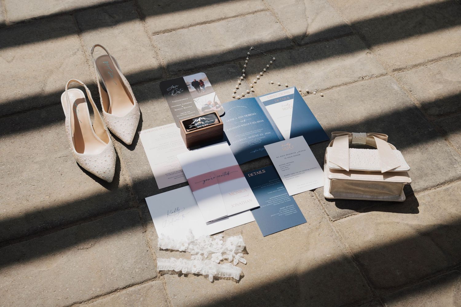 Elegant wedding flat lay with ivory heels, clutch, blue stationery suite, ring box, and jewelry on stone pavement.