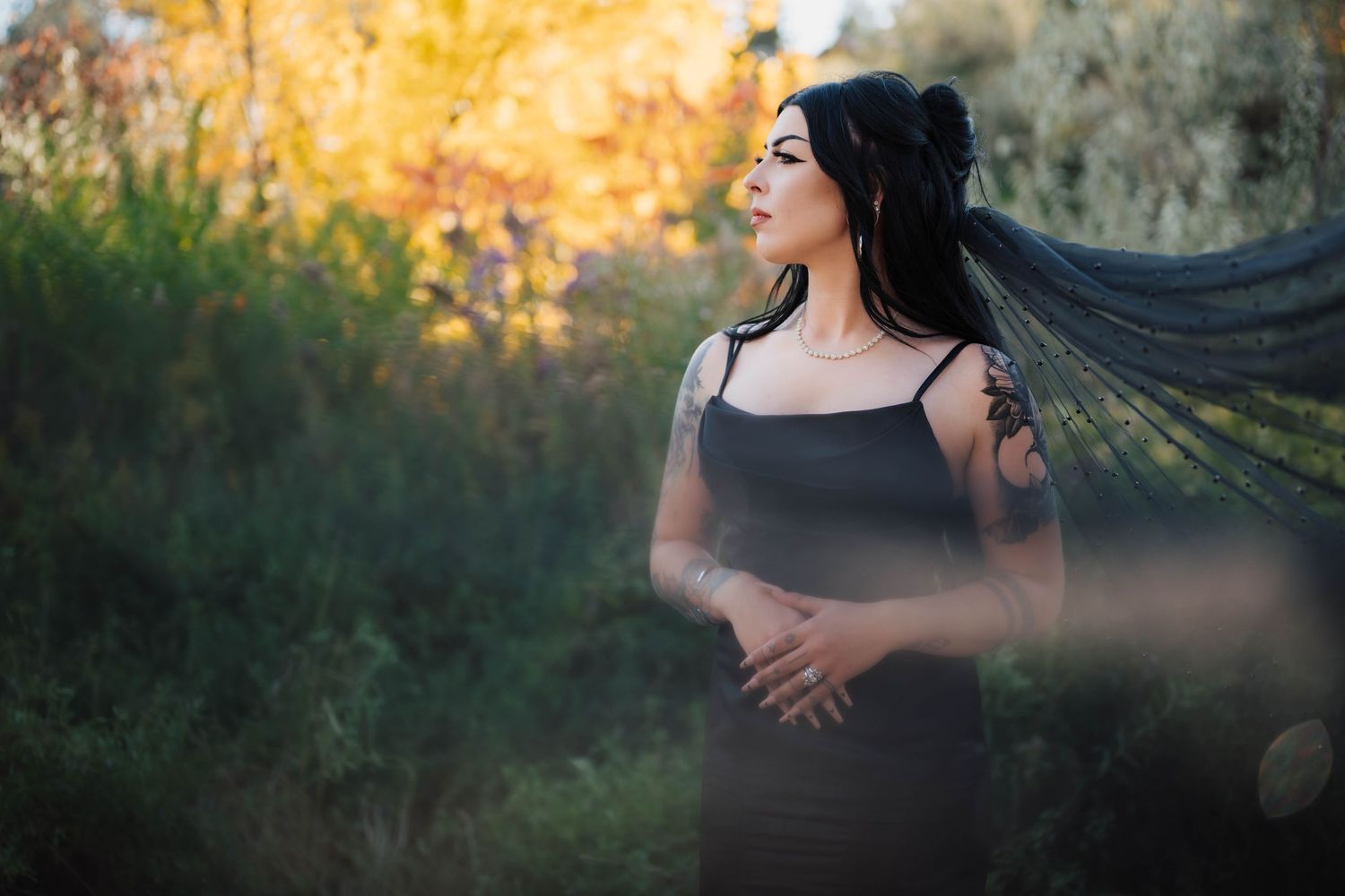 Woman in black dress with flowing veil stands outdoors among lush greenery in golden hour light, showcasing arm tattoos.