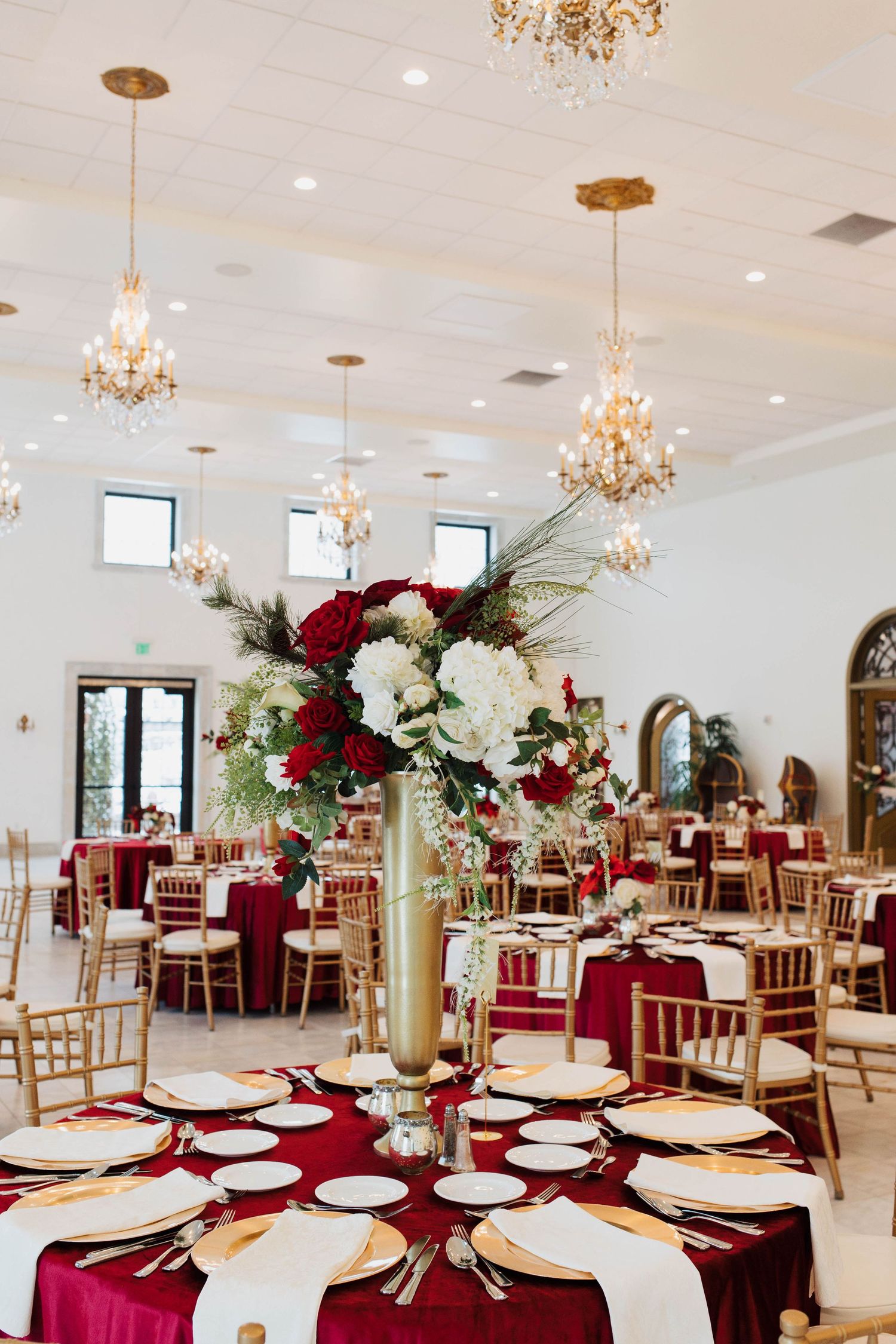Elegant wedding reception hall with crimson velvet tablecloths, gold chiavari chairs, tall floral centerpieces, and crystal chandeliers.