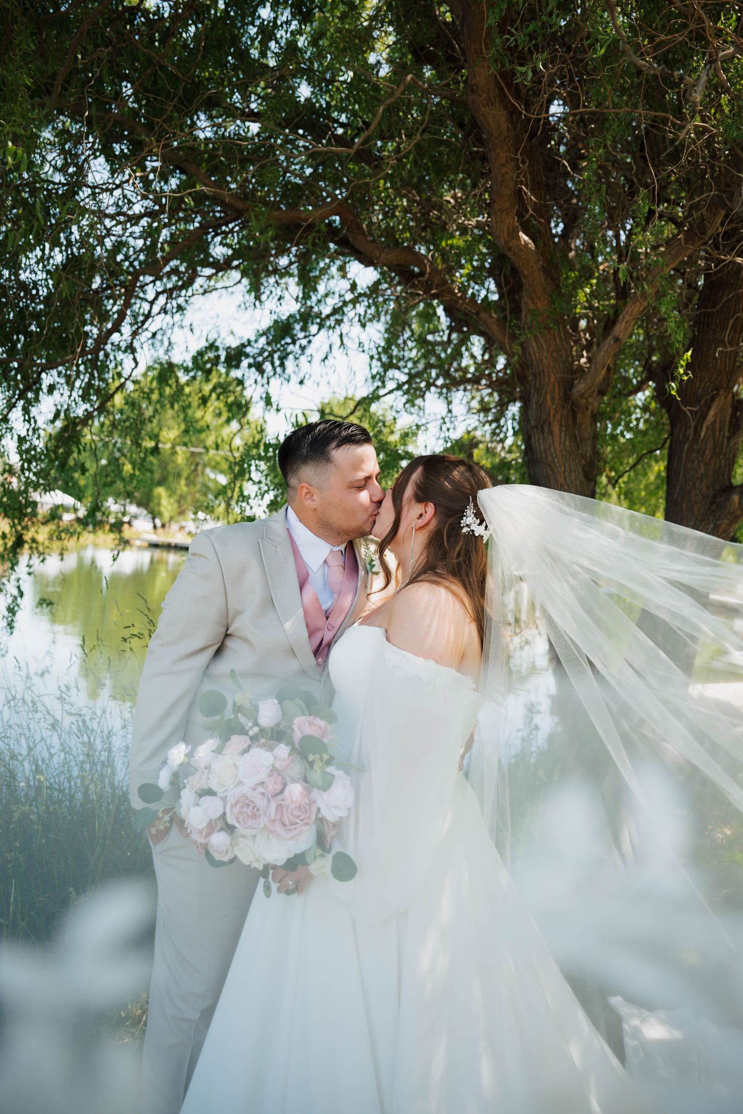 Bride and groom share a kiss under a tree by a lake, veil flowing, as bride holds a blush pink bouquet.