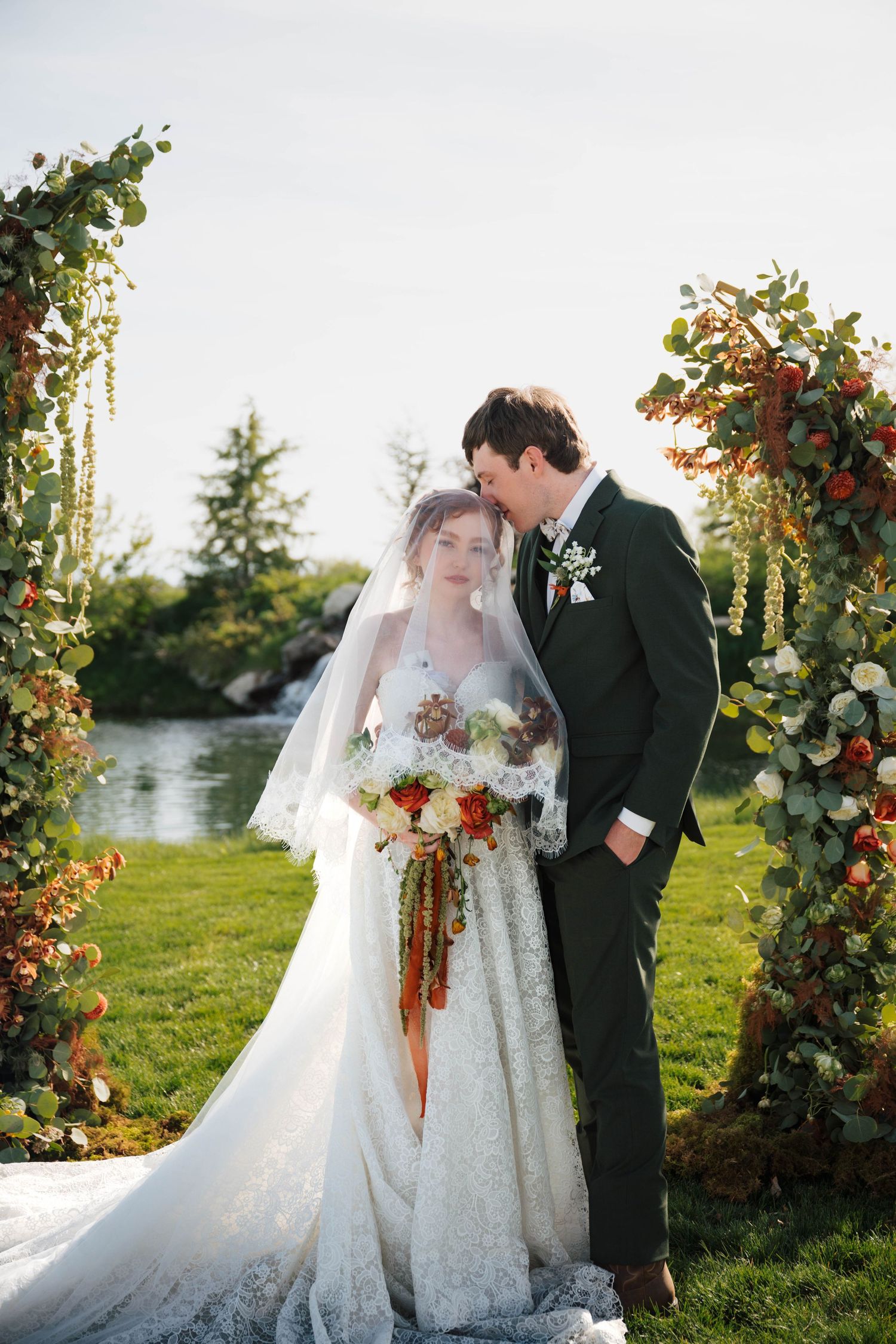 Bride and groom pose under floral arch with autumn blooms by a lake, bride holding cascading bouquet in lace gown.