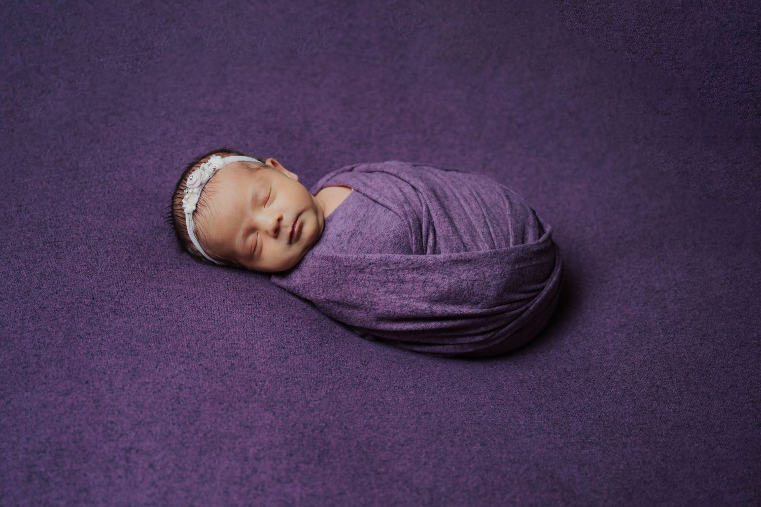 Newborn baby swaddled in purple wrap with floral headband, sleeping peacefully on purple background.