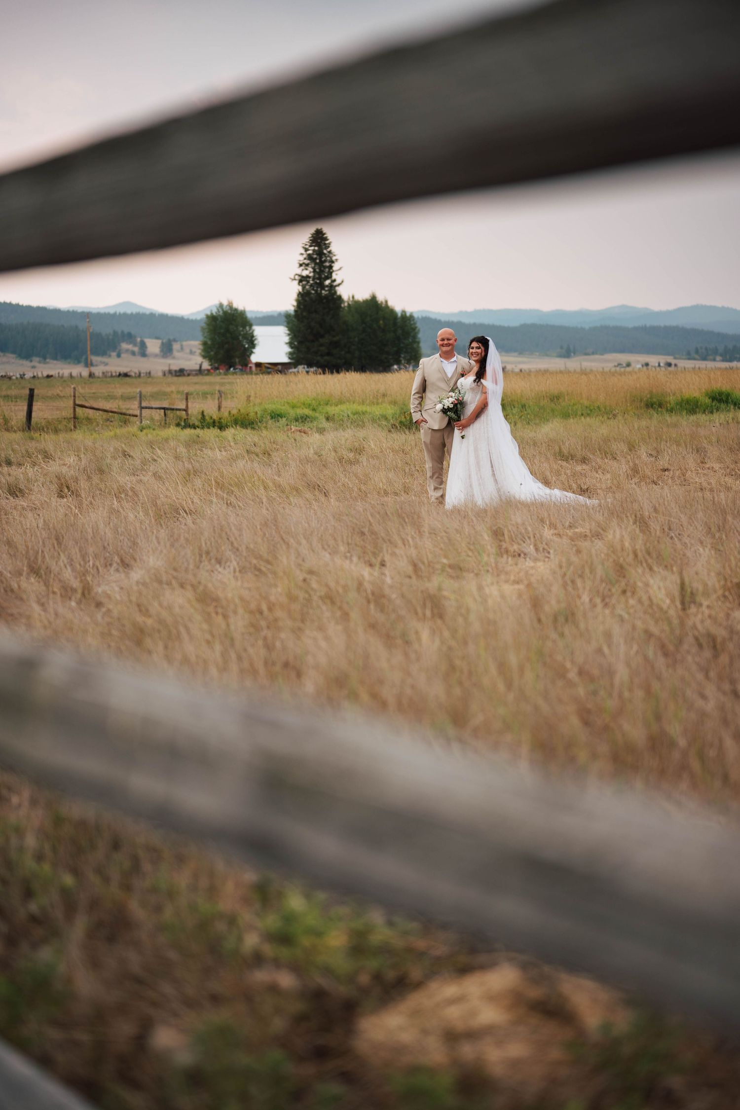 A wedding couple stands in a golden field framed by a rustic wooden fence.