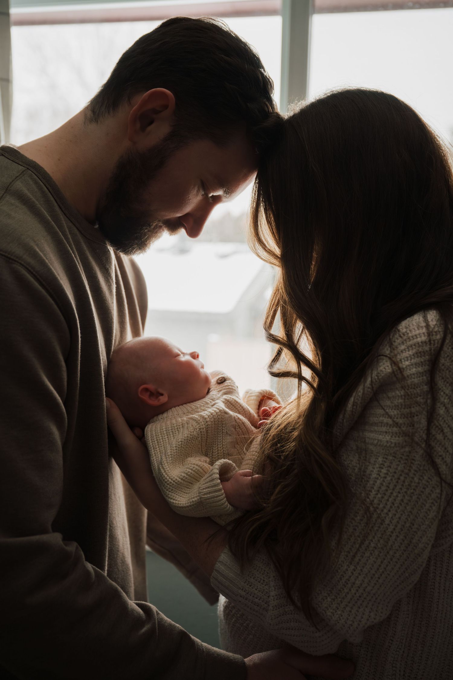 Silhouetted family moment showing parents tenderly holding baby near window.