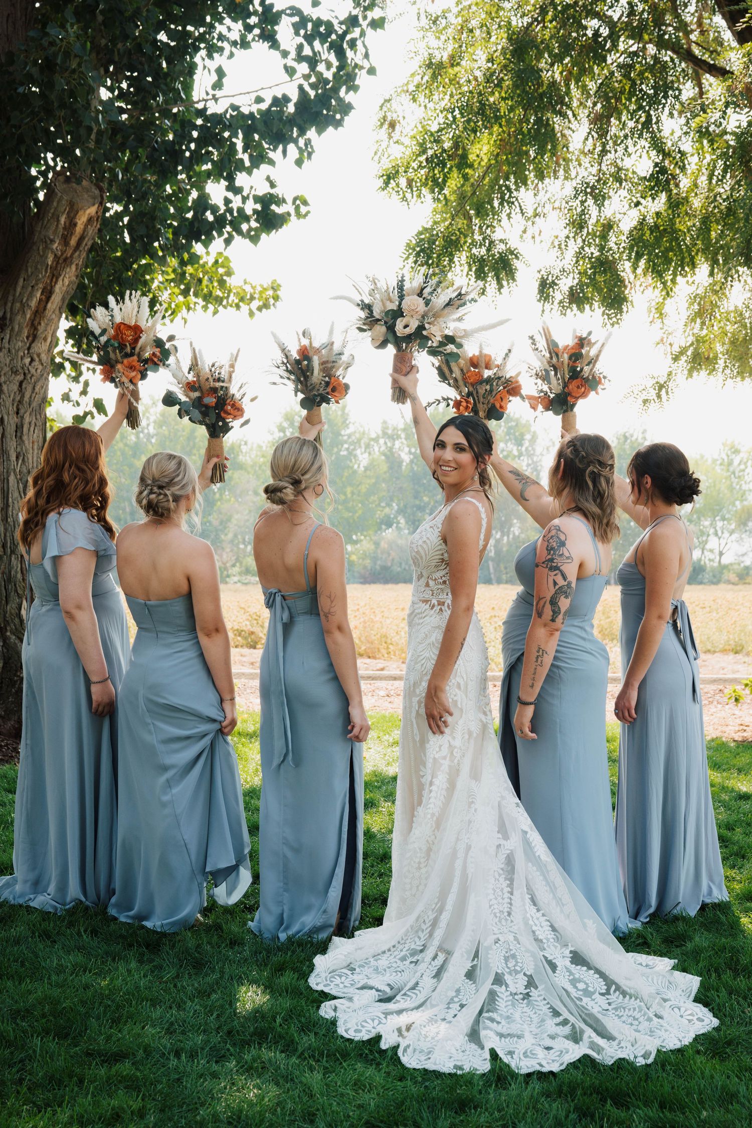 Bridesmaids in dusty blue dresses standing in a row with the bride in a lace gown outdoors.