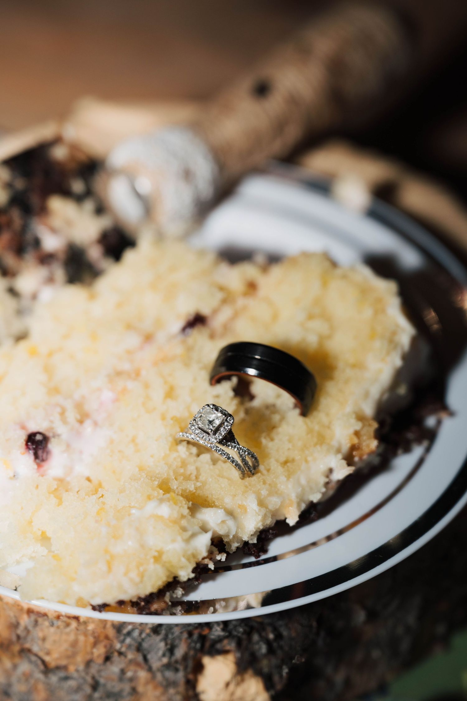 Wedding rings, a black band and diamond engagement ring, rest on a slice of white cake with berries on a rustic wooden surface.