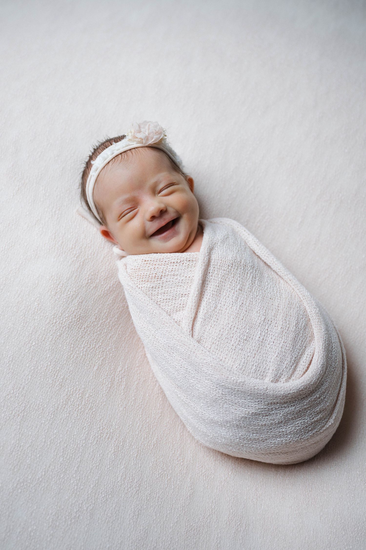 Smiling newborn baby swaddled in white wrap with floral headband lying on soft white background.