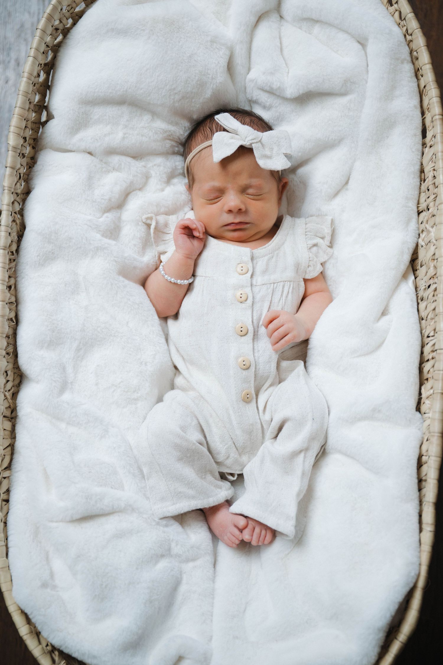 Sleeping newborn baby girl in white romper and bow headband resting peacefully in a wicker Moses basket.
