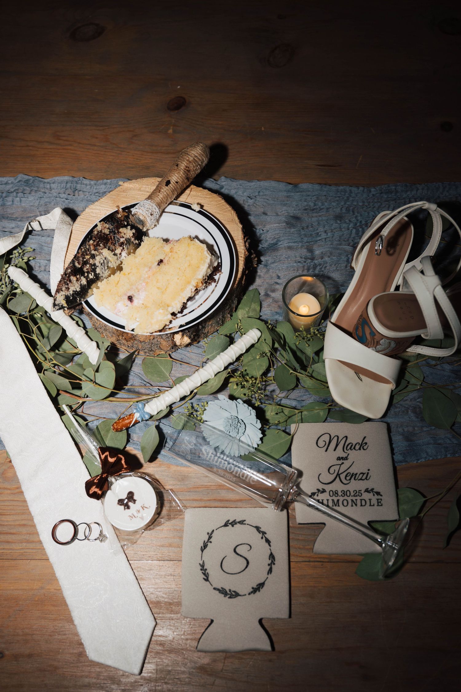 Wedding details flatlay with rings, cards, and eucalyptus leaves on dark wooden surface.